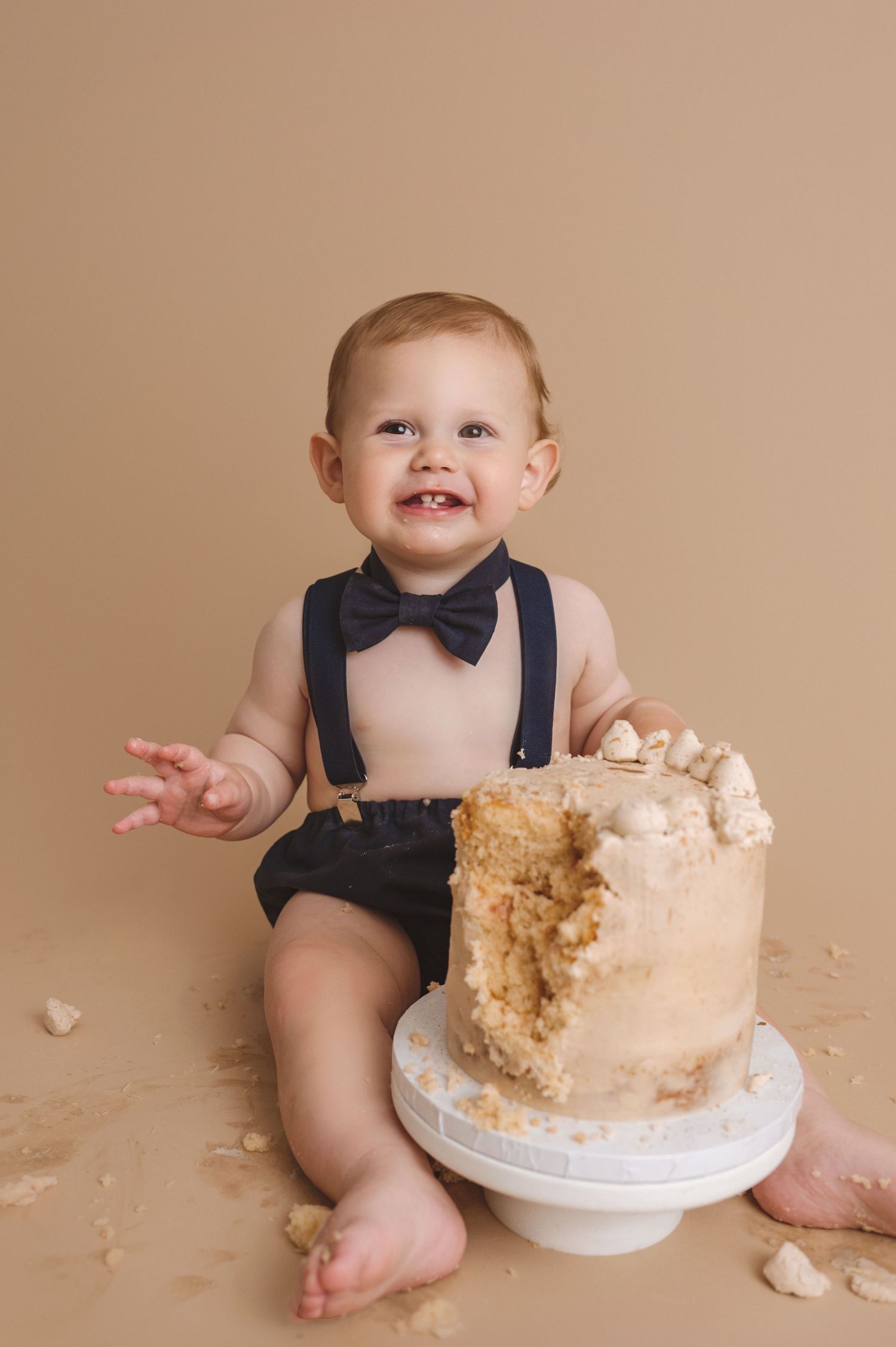 Sweet cake smash photography session featuring a baby in black suspenders and bow tie sitting beside a partially destroyed cake.