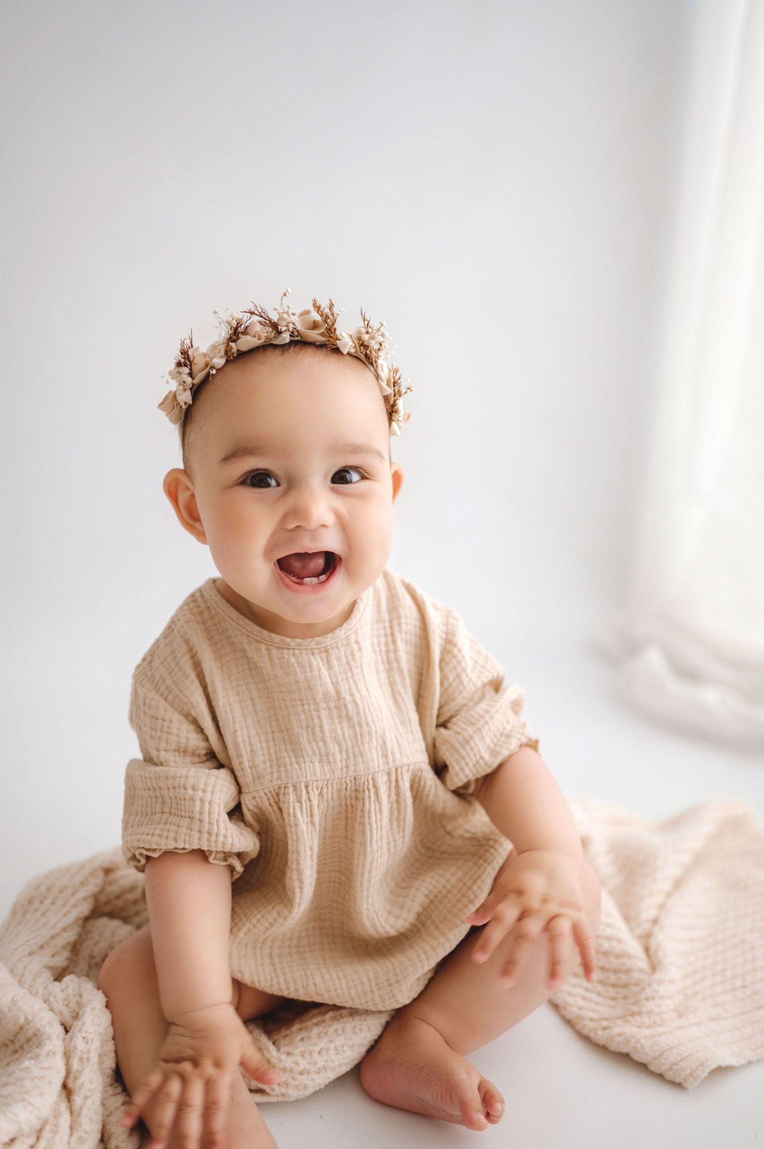 Baby wearing cream dress and floral crown sitting on floor smiling joyfully against white wall.