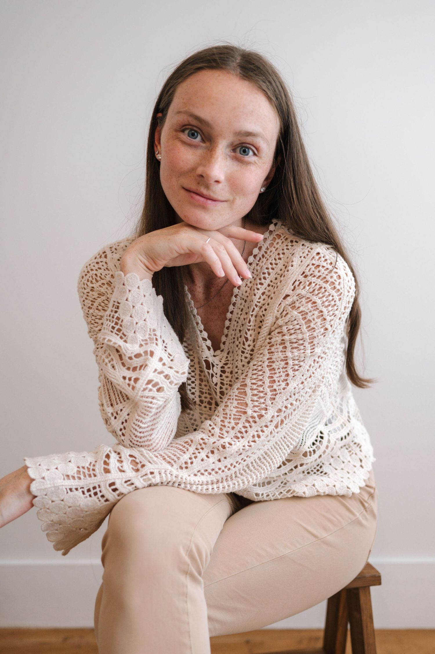 A person in a cream-colored crocheted sweater poses thoughtfully against a white wall in a minimalist setting.
