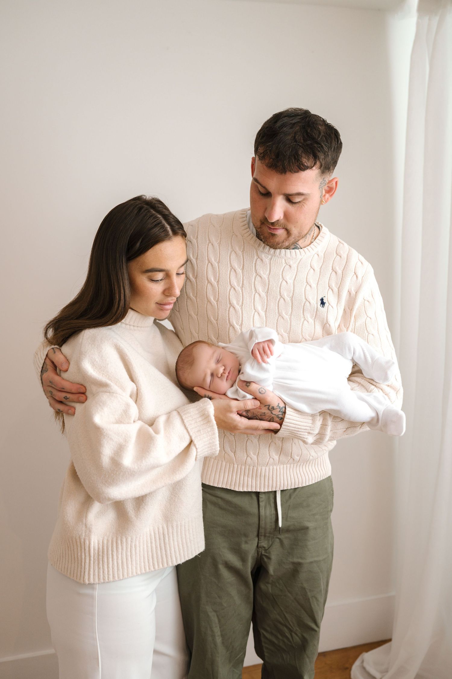 Young couple in cream sweaters tenderly gazing at their sleeping newborn baby in a bright white room.