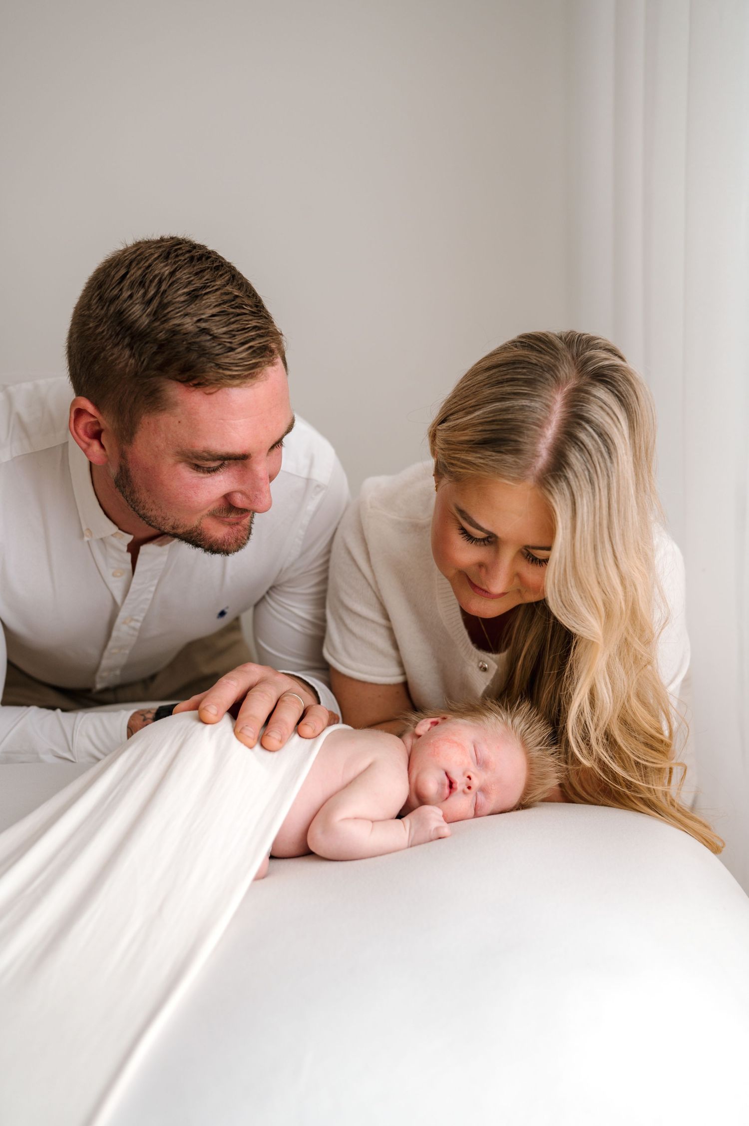 Parents lovingly gaze at their sleeping newborn baby wrapped in white cloth during a professional newborn photo session.