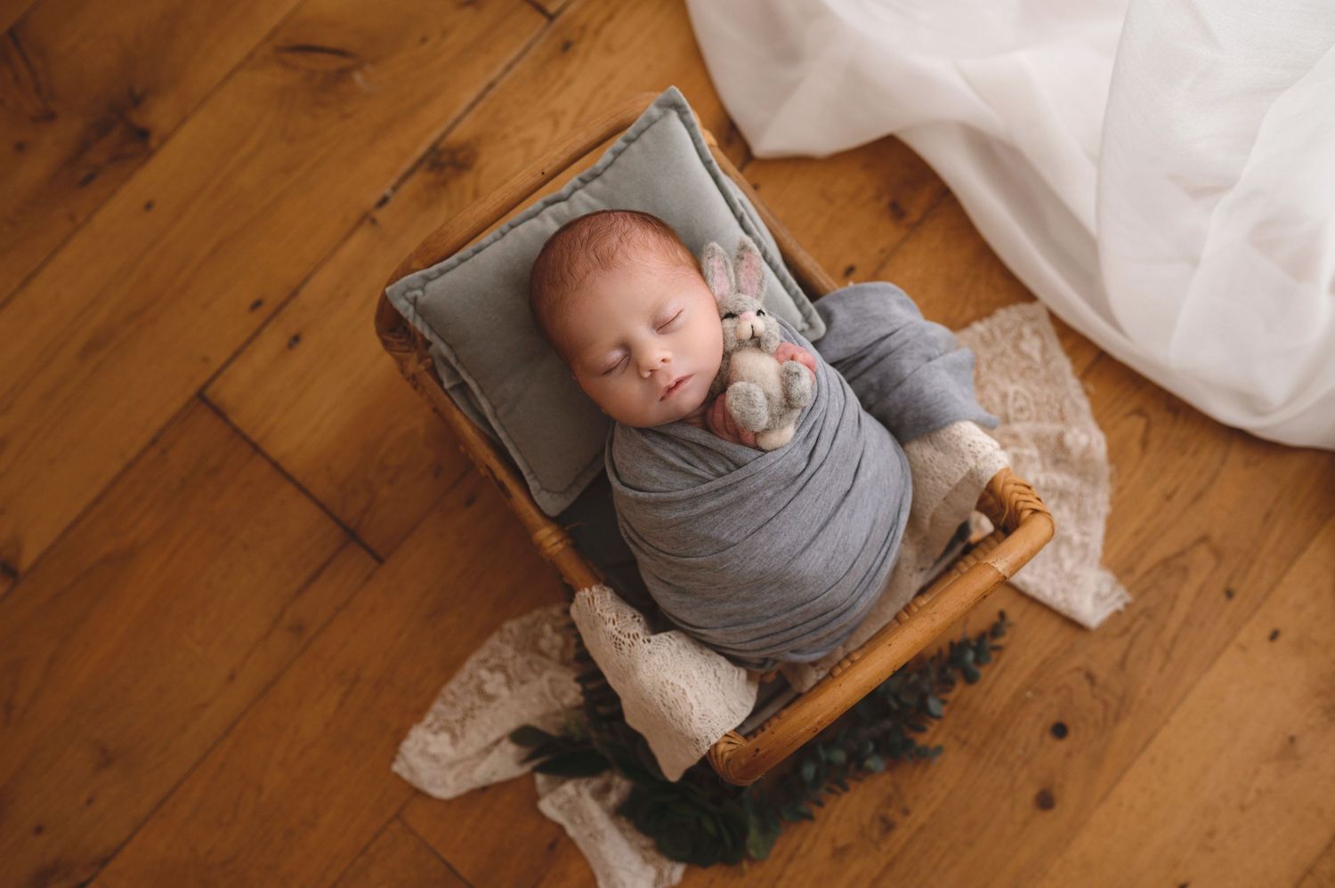 Baby wrapped in gray swaddle lying in vintage wooden drawer on hardwood floor with lace fabric.