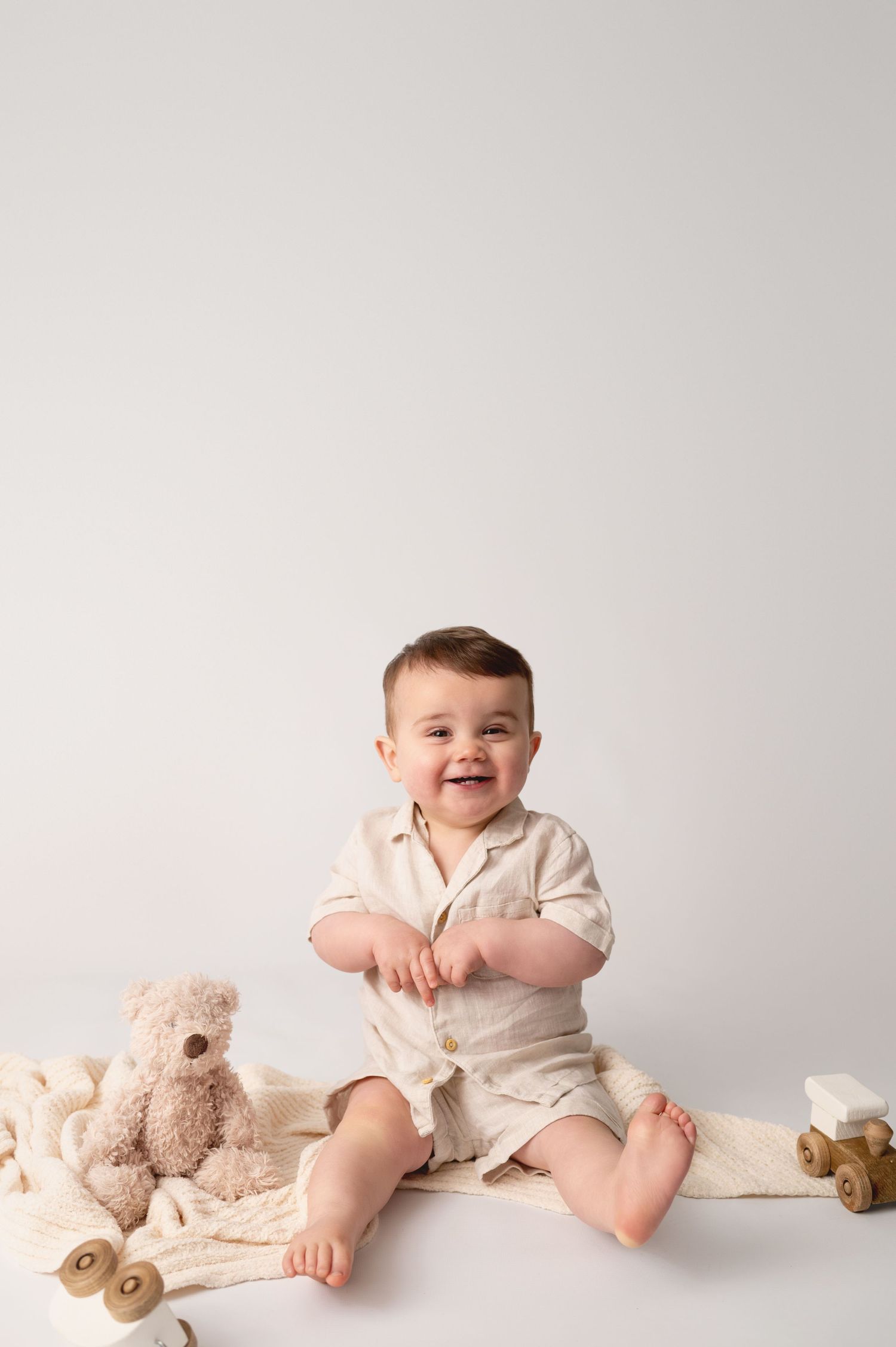 A happy baby in white clothing sits on a cream blanket with plush toys and wooden decor.