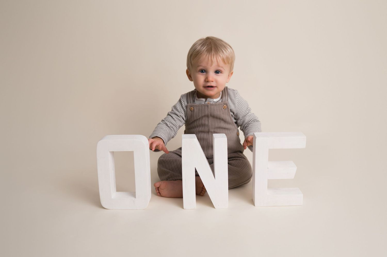 A toddler in a gray outfit sits with large white decorative letters spelling ONE on a neutral backdrop.