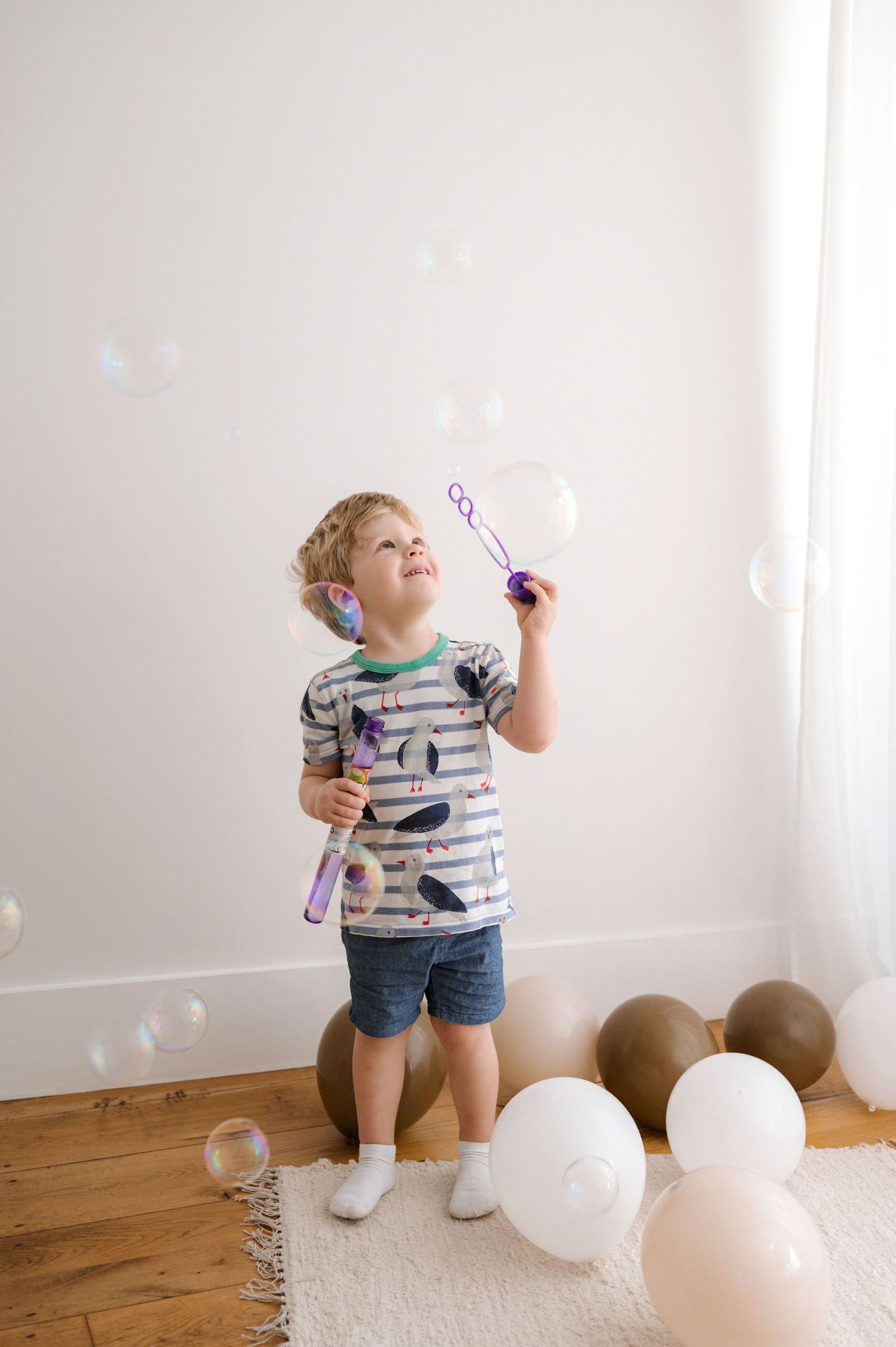 A child in a striped shirt blows bubbles while standing on a wooden floor with white balloons nearby.