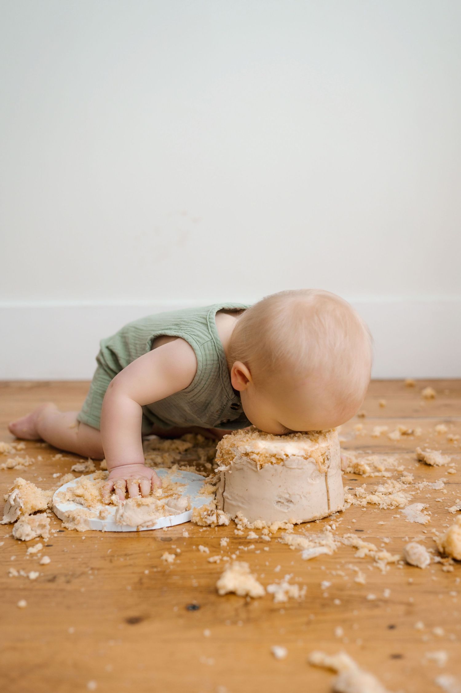 Baby in mint green romper leaning over to eat cake off wooden floor during messy cake smash photo session.