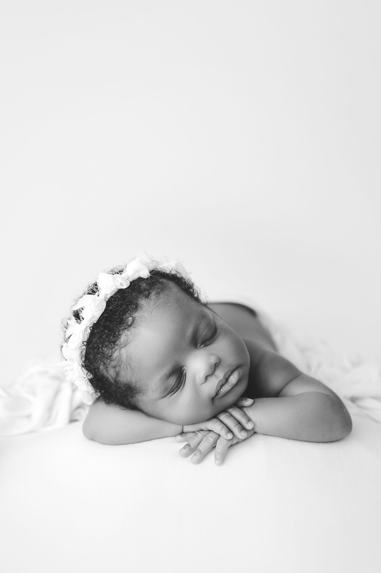 A sleeping baby wearing a small crown rests peacefully on soft white bedding in black and white style photography.
