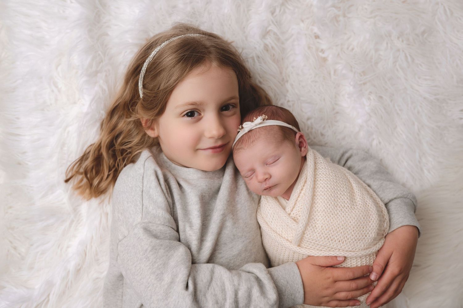 A young child in gray sweater lovingly embraces a sleeping newborn swaddled in cream-colored fabric on white background.