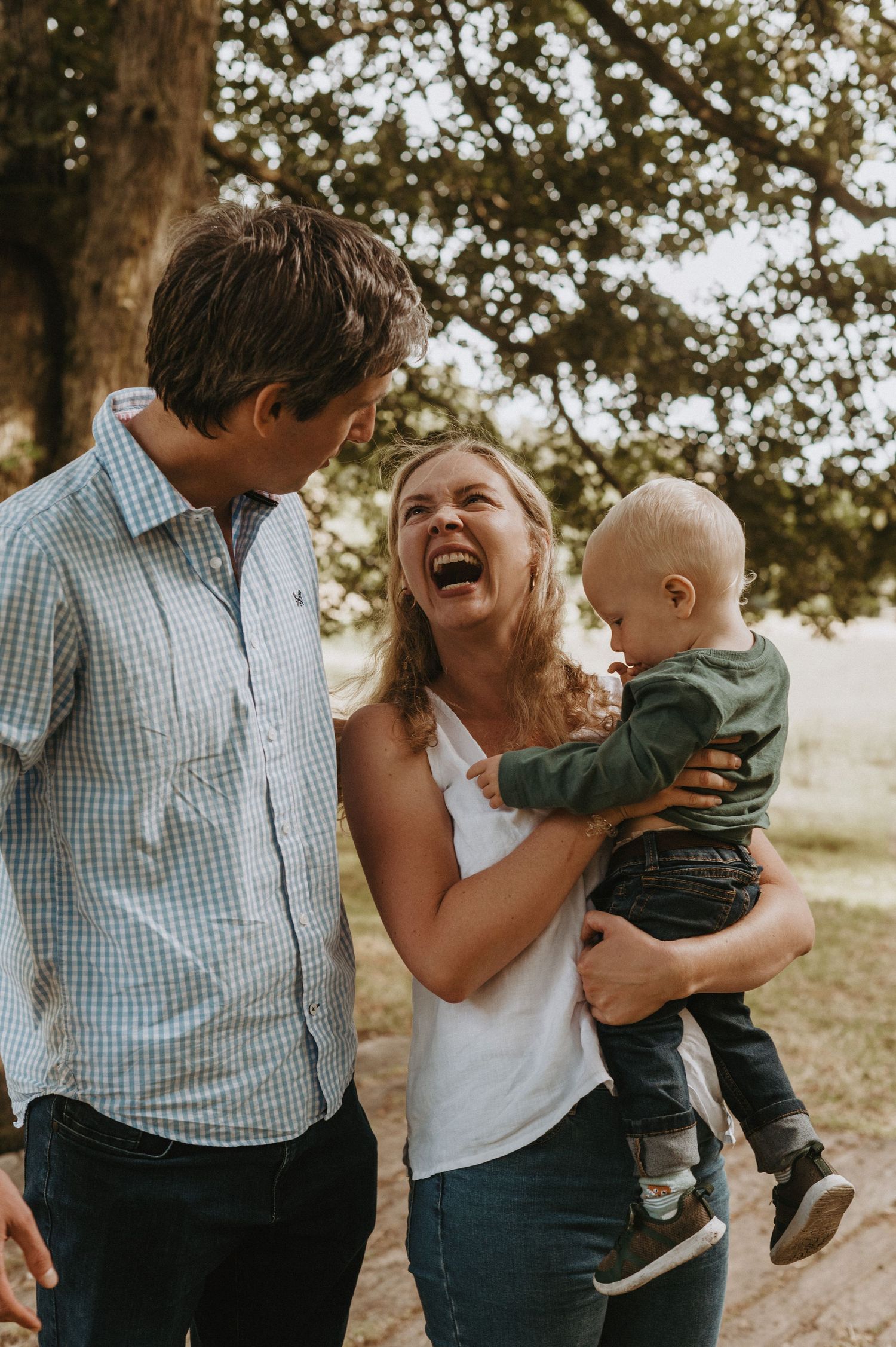 Family share a candid joyful moment outdoors beneath a tree in warm sunlight.