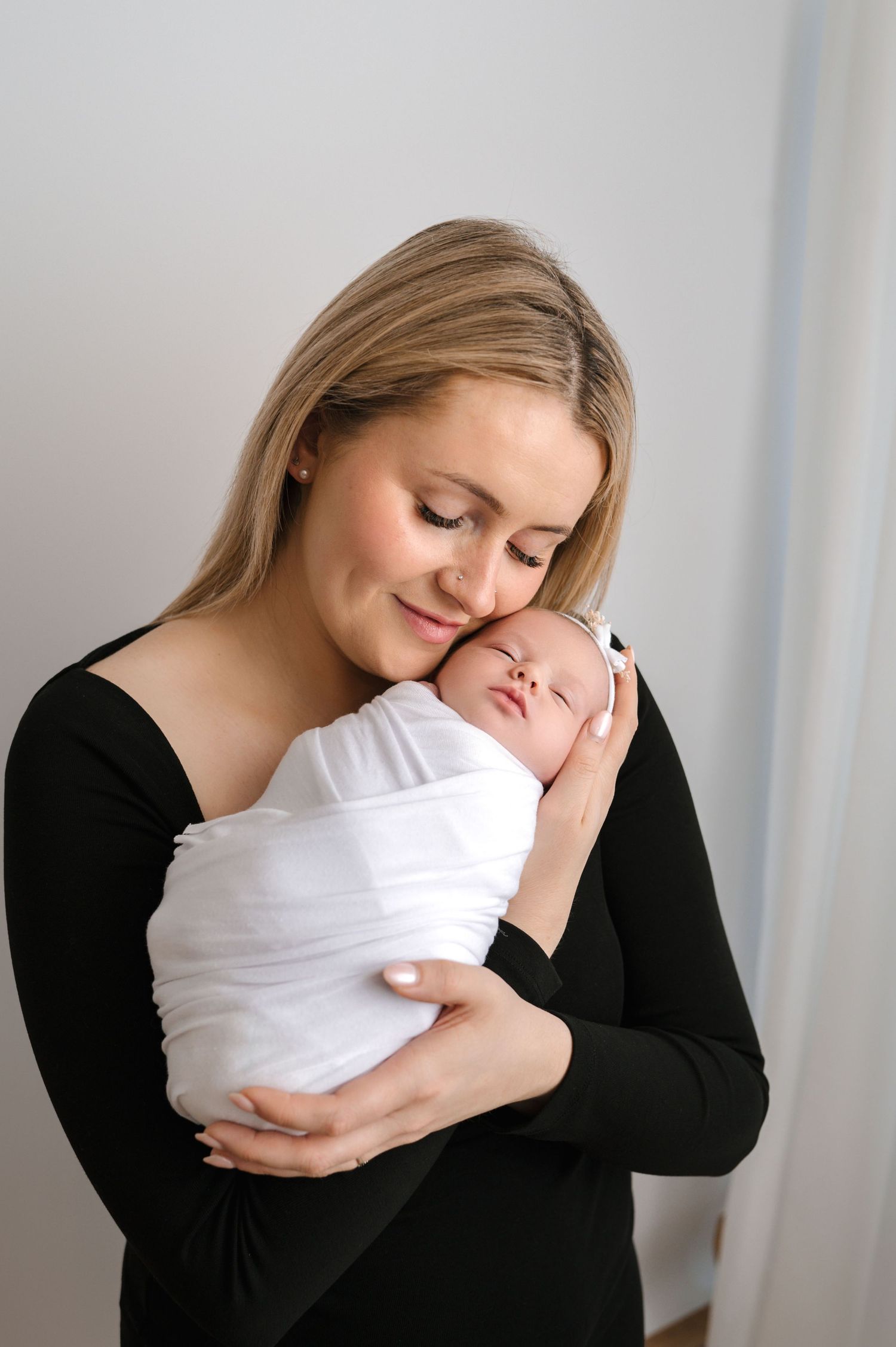 A tender moment captured as a mother in black embraces her swaddled newborn baby against a light background.