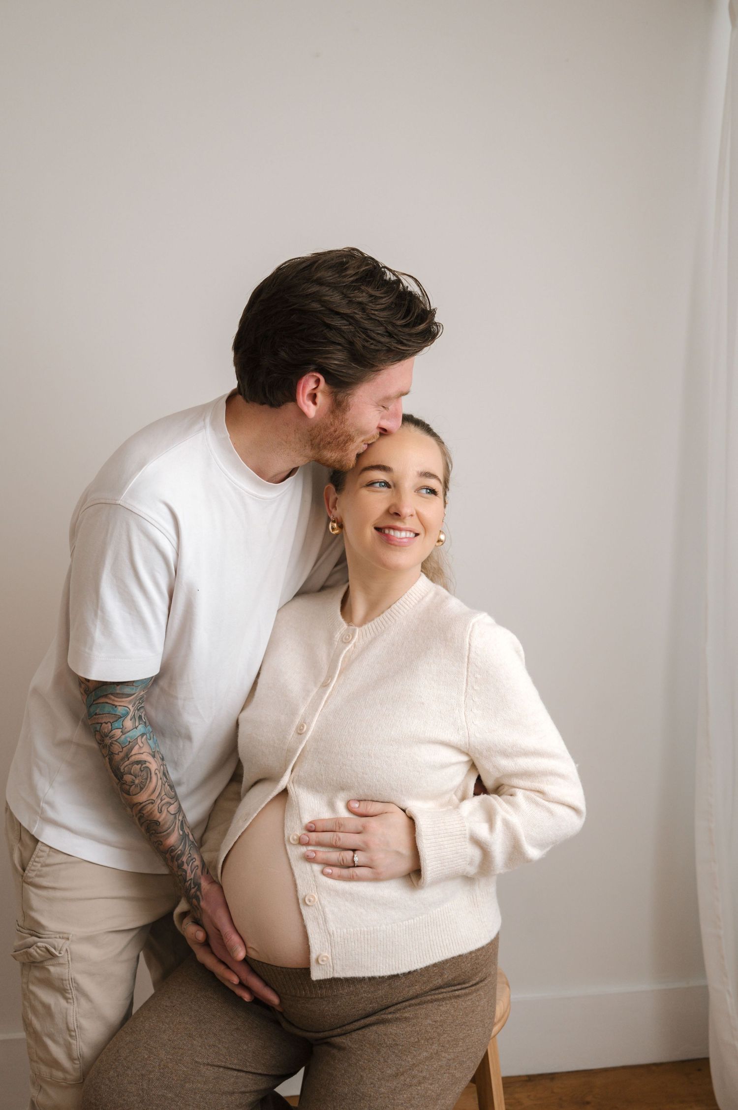 A couple in white and beige clothing embraces tenderly in a minimalist studio setting against a light background.
