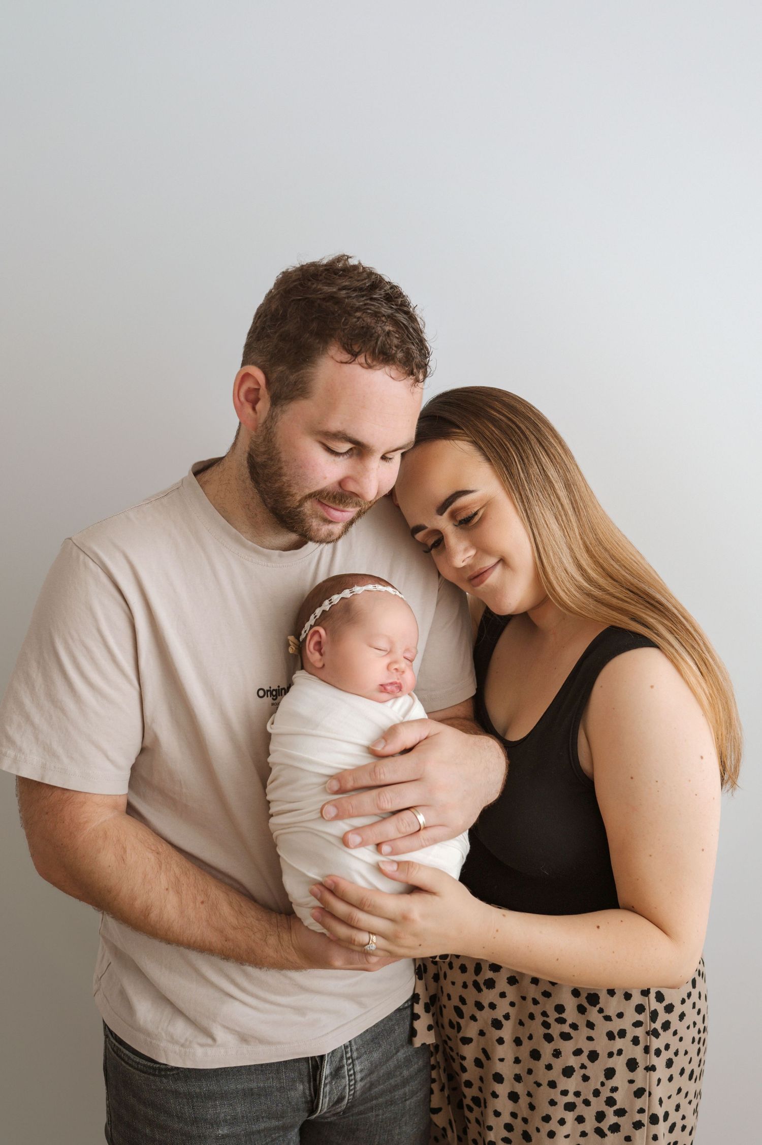 New parents tenderly hold their swaddled newborn baby girl wearing a white headband in a studio portrait.