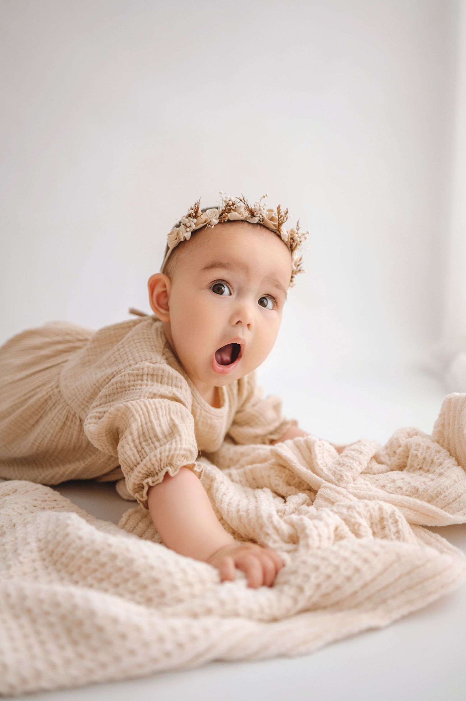 A baby in a cream-colored outfit lies on a textured blanket while looking up with an expressive face.