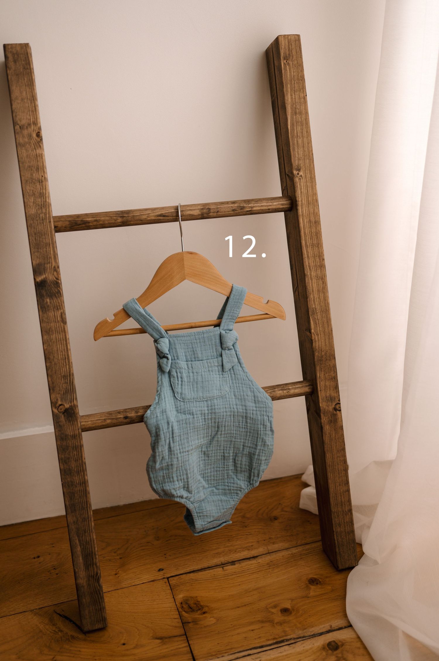 A light blue baby romper hangs on wooden hangers against a rustic wooden ladder in a minimalist setting.
