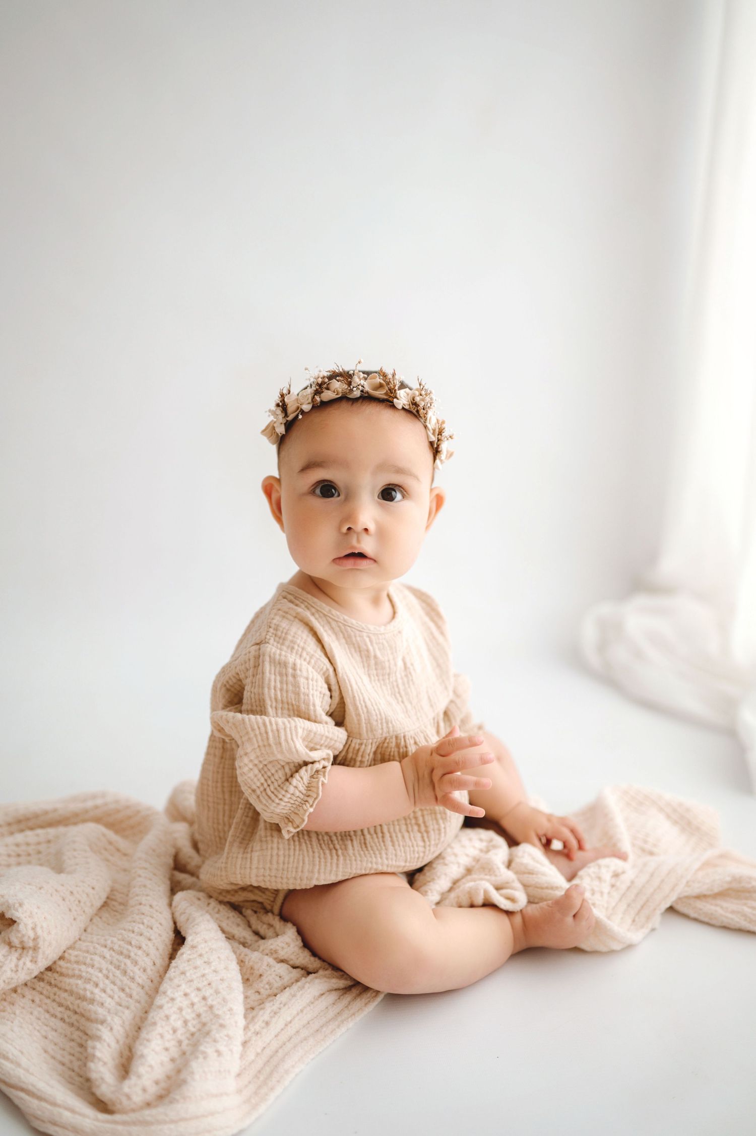 Adorable baby girl sitting on a cream waffle blanket wearing a beige knit romper and floral crown headband.