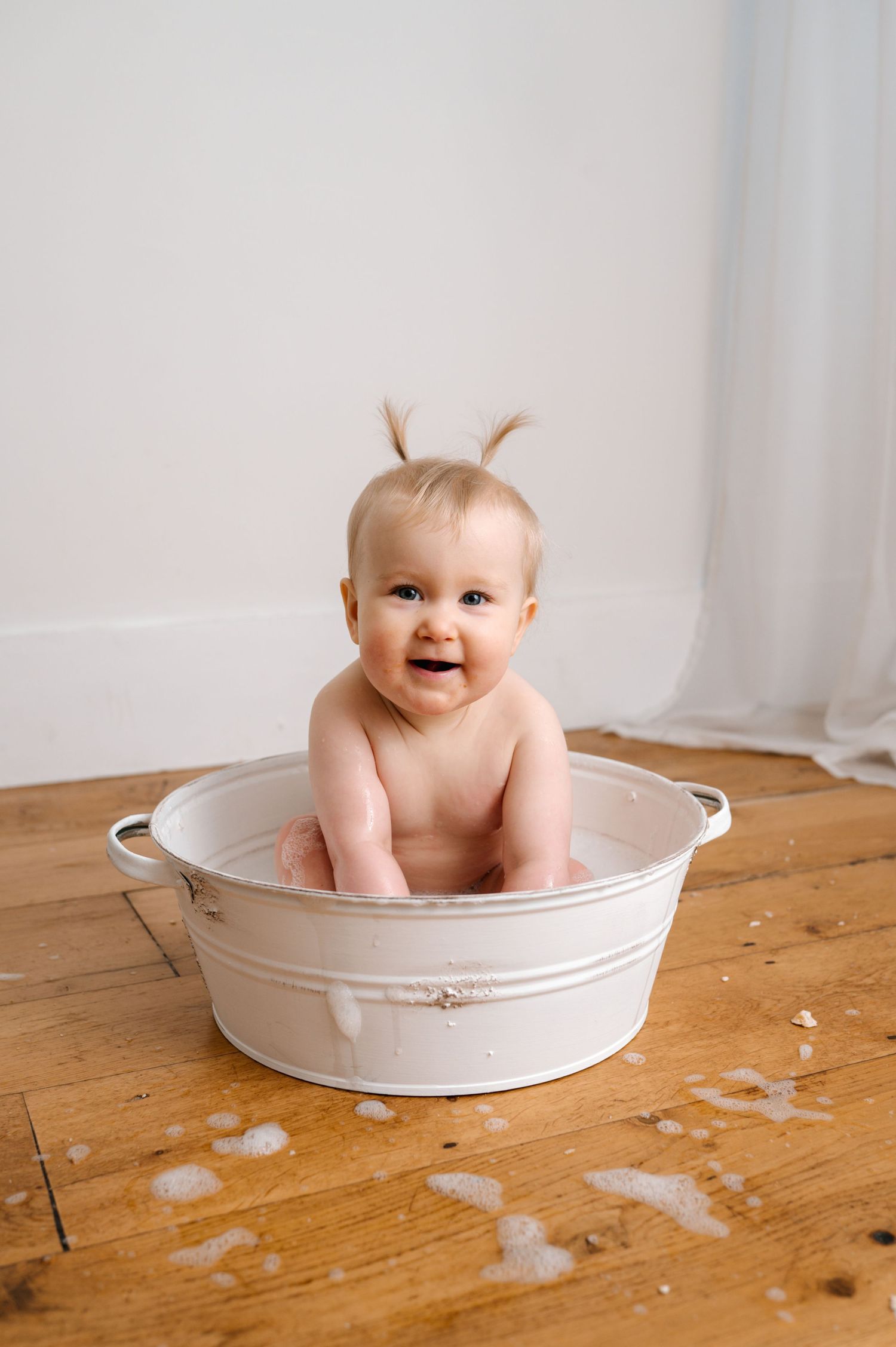 Smiling baby sitting in a white metal tub on a wooden floor with soap bubbles around it.