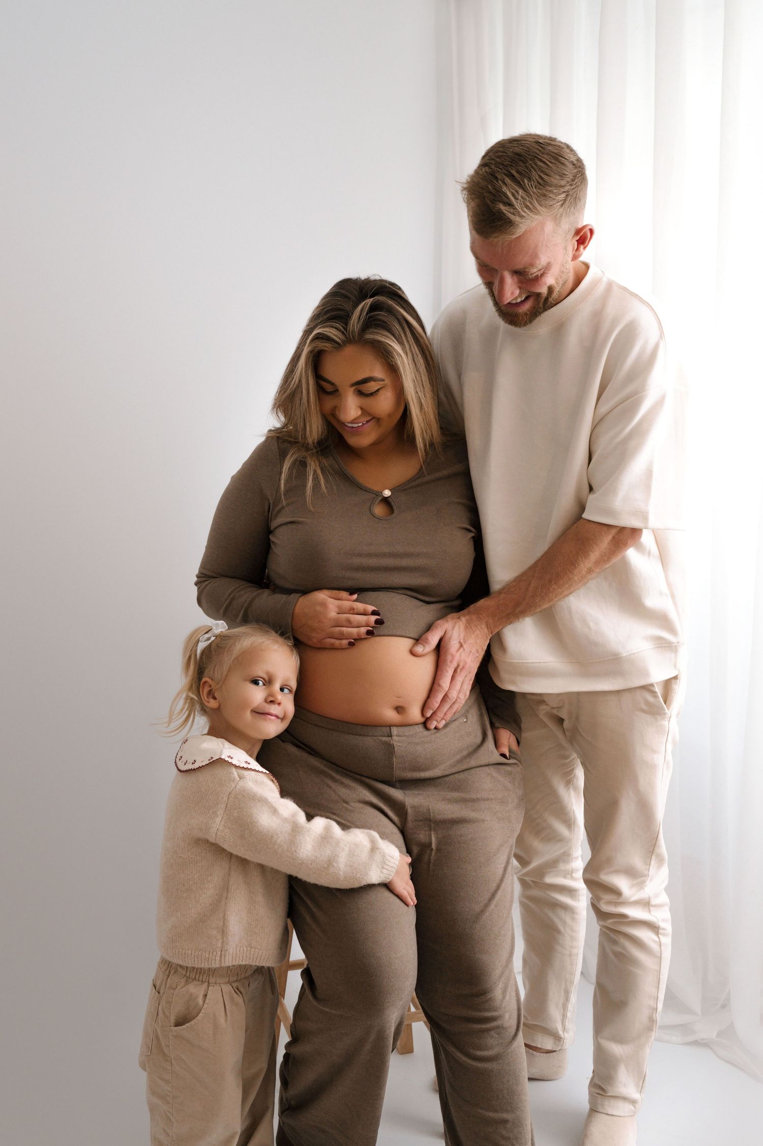 A pregnant woman in brown outfit poses with her smiling husband and young daughter hugging her baby bump.