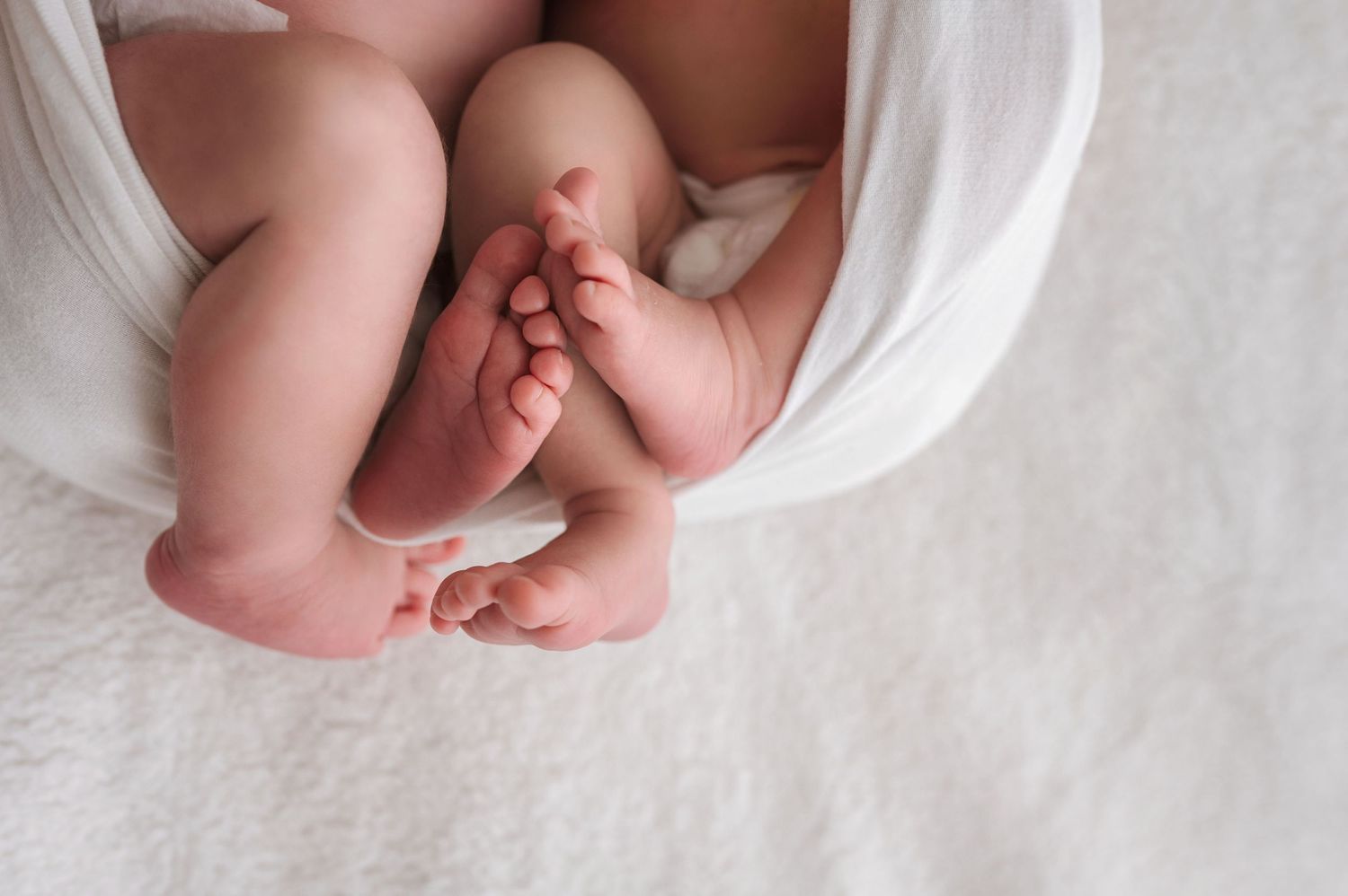 Close-up of newborn baby feet and legs wrapped in a soft white blanket on a white textured surface.