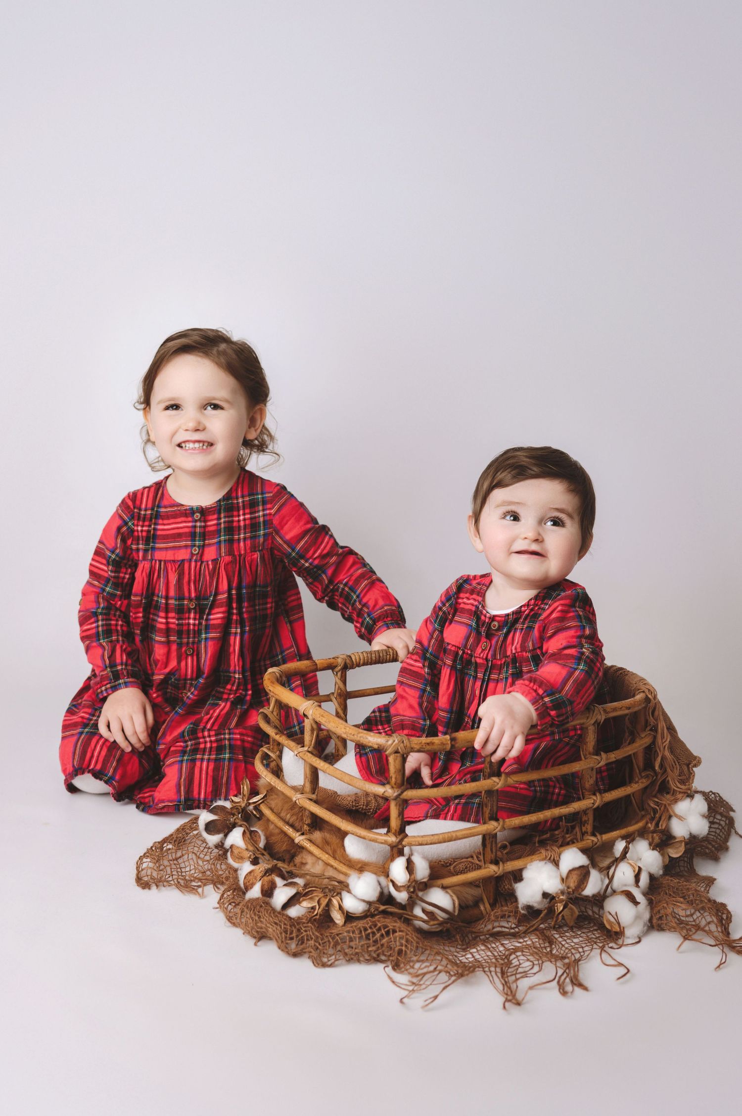 Two children in matching red plaid dresses sit with a rustic wooden basket and cotton branches on a light background.