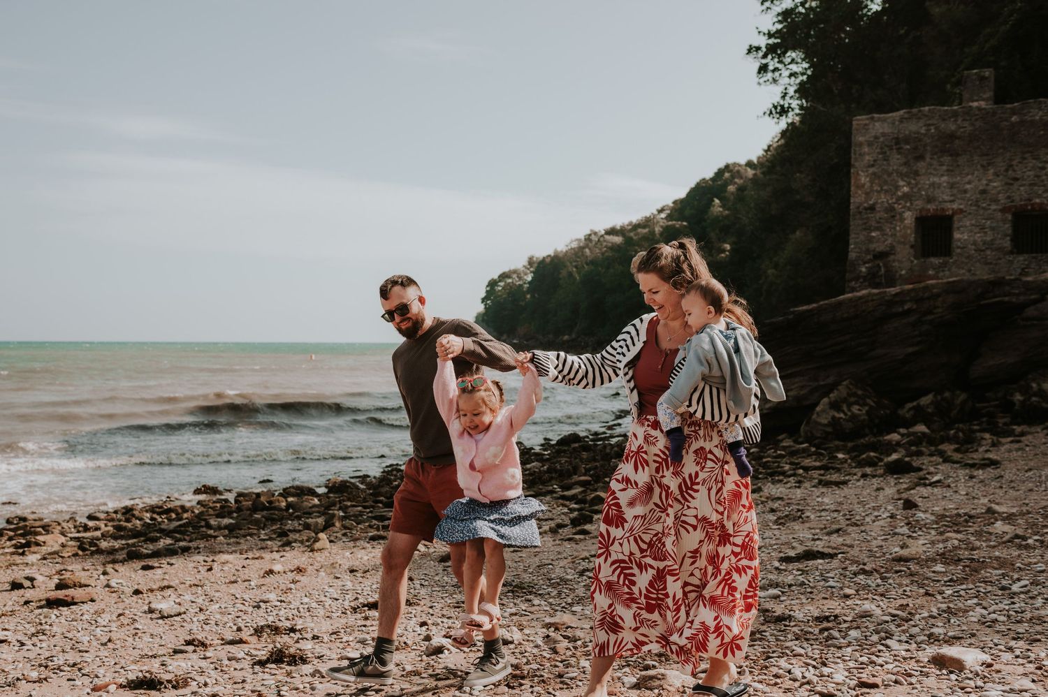 Family walking and dancing together on a rocky beach against the backdrop of an old stone structure by the ocean.