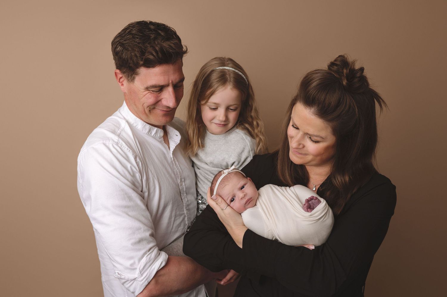 A family of four gathers for a studio portrait against a warm brown backdrop.