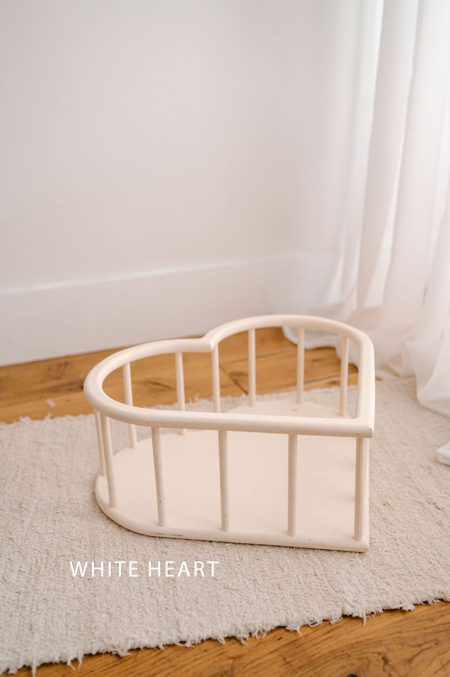 White heart-shaped wooden storage basket on beige carpet and hardwood floor near sheer curtains.
