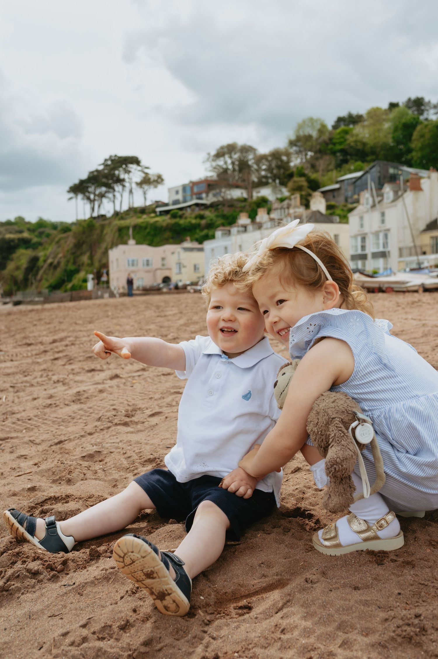 Two young children sit on a sandy beach, smiling and pointing, with coastal village and green hills behind.