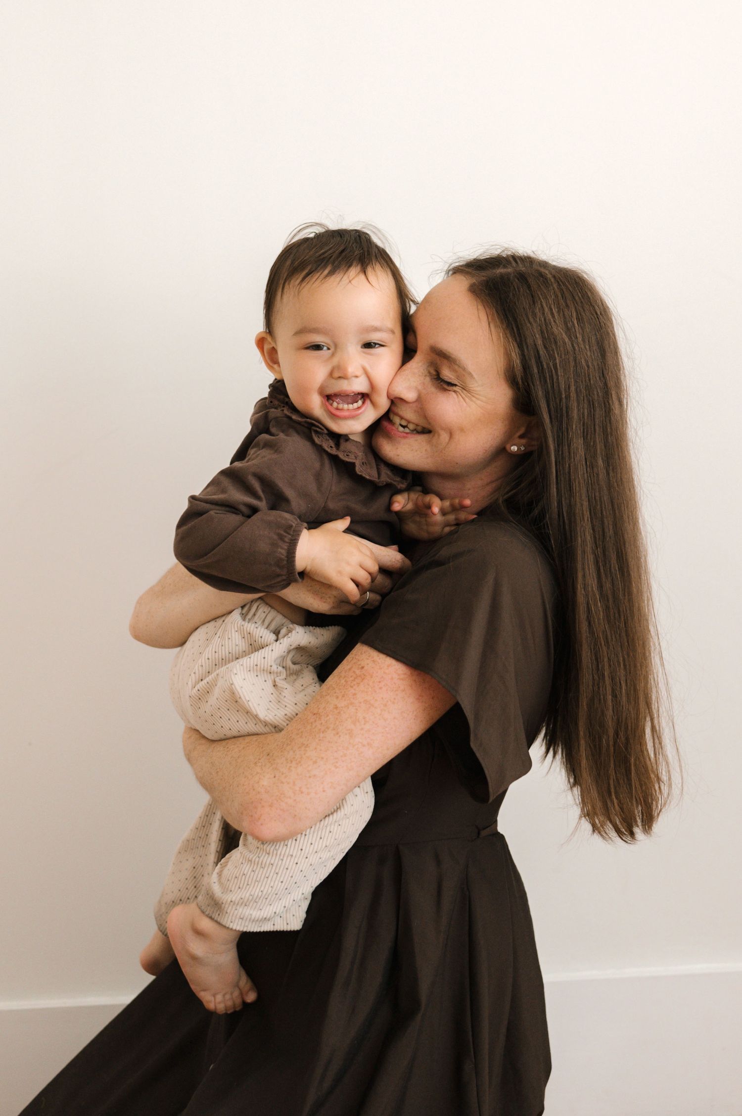 A smiling woman in a dark dress holds a laughing baby in a brown top against a white background.