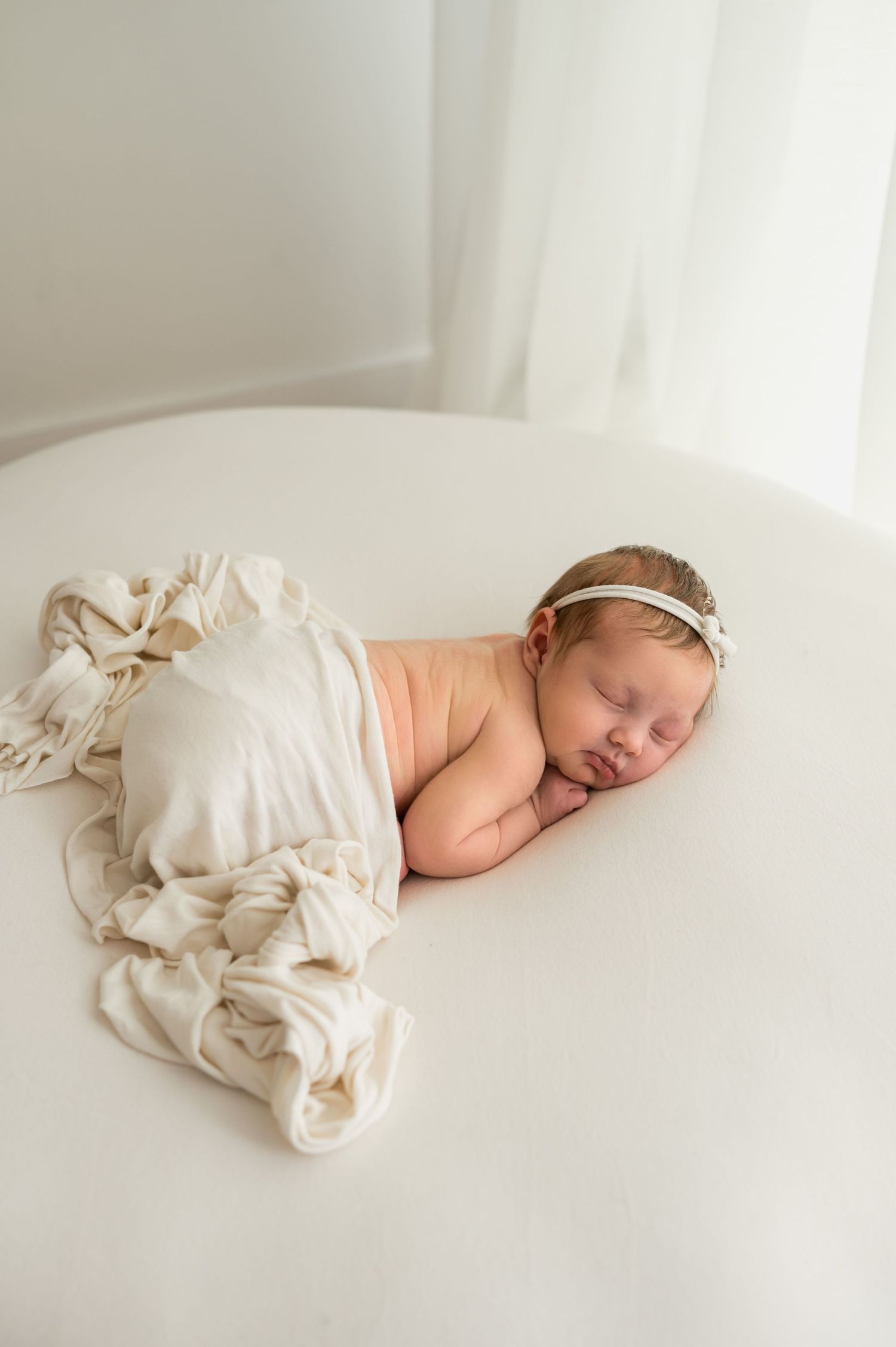 Sleeping newborn baby girl with floral headband on white round surface with cream fringe wrap.