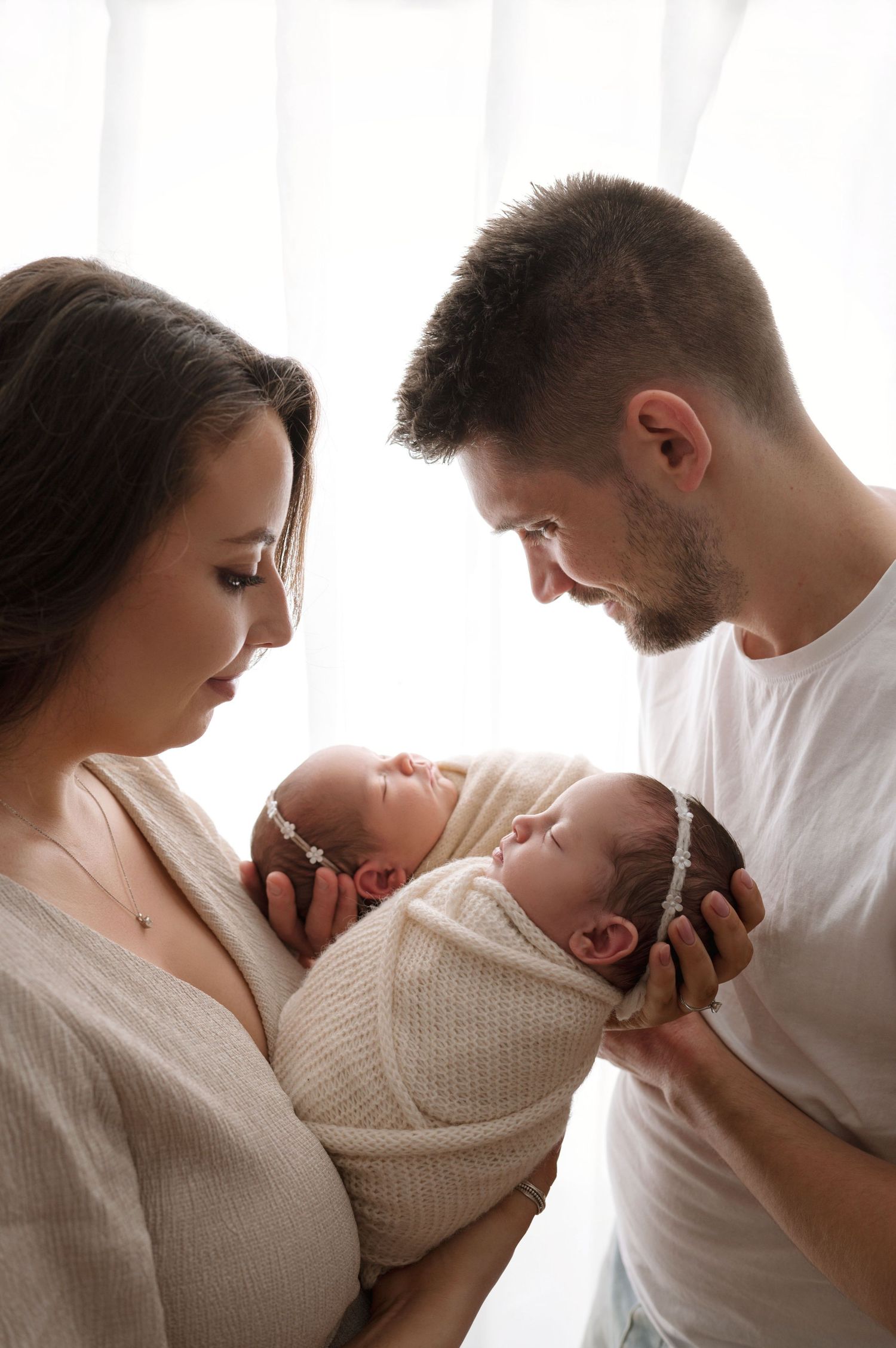 Parents in white outfits gaze lovingly at their twin newborns wrapped in cream-colored knit blankets against soft lighting.