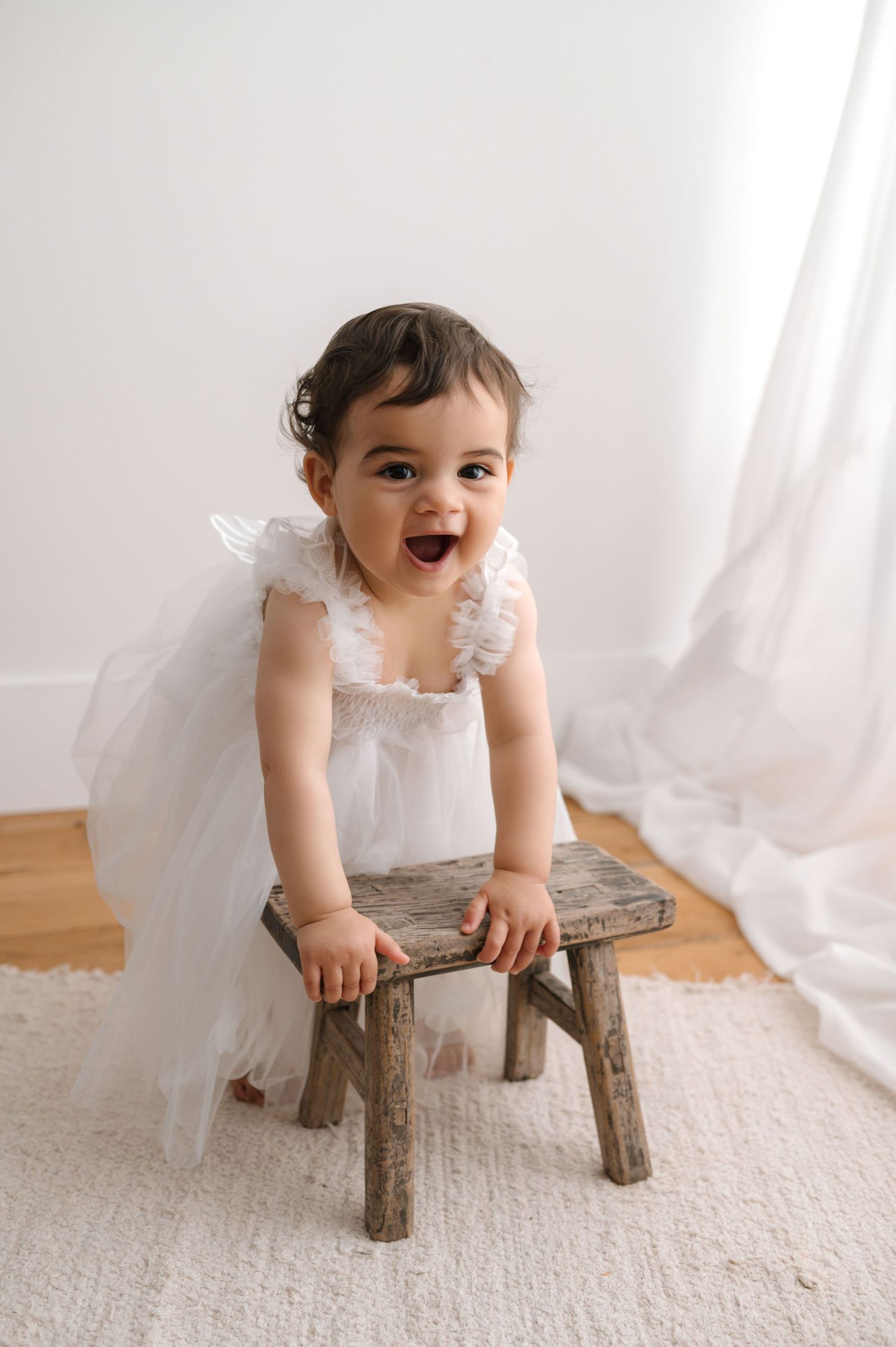 A cheerful baby in a white lace dress stands while holding onto a rustic wooden stool against a light background.