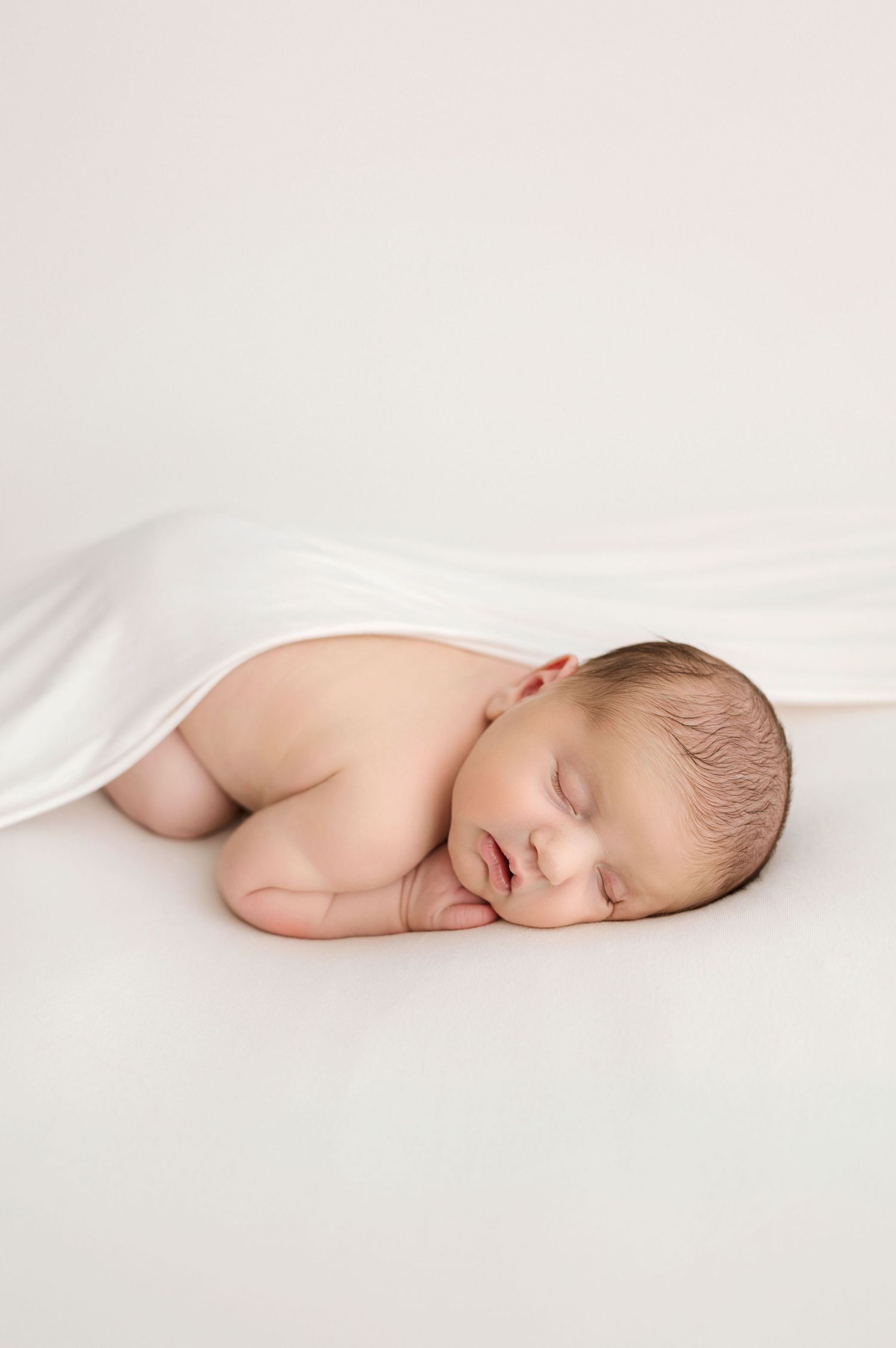 A sleeping newborn baby lies peacefully under white fabric in a minimalist portrait photograph.