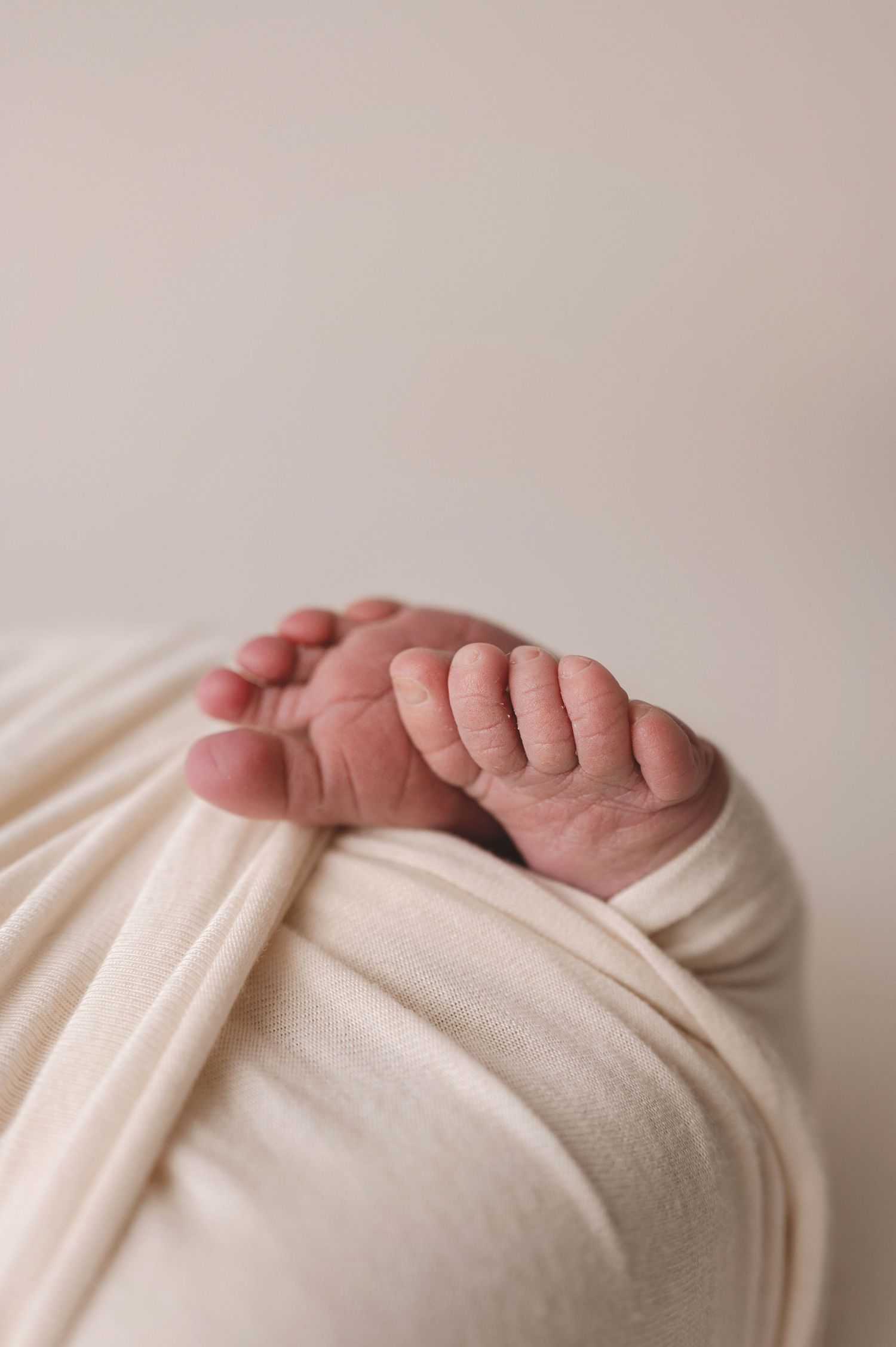 Tiny newborn baby toes peek out from soft white swaddling blanket against neutral background.