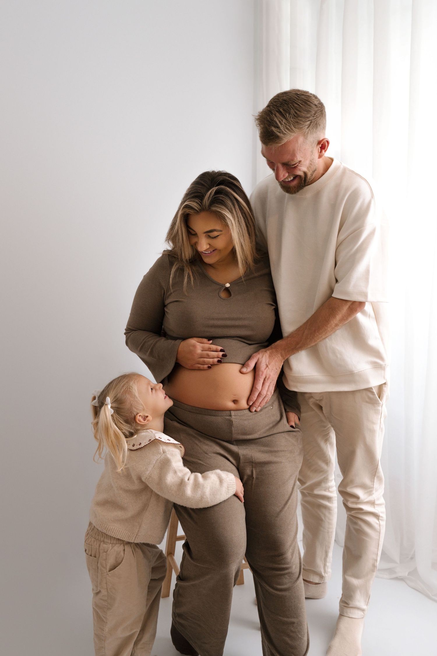 A family in neutral-toned clothing shares tender moments together during a maternity photoshoot in a bright room.