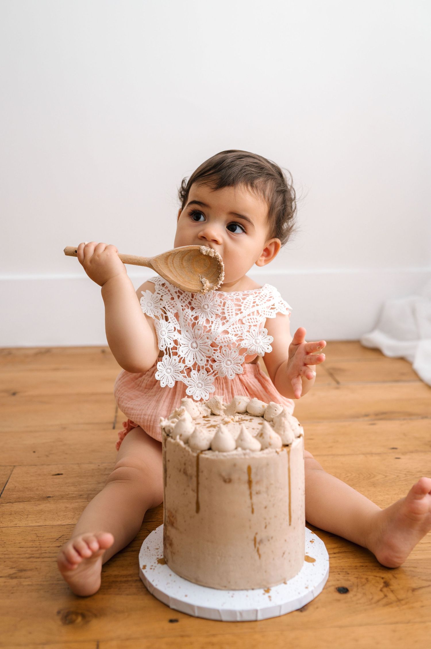 A baby in a lace dress sits on a wooden floor smearing cake frosting while enjoying a chocolate birthday cake smash session.