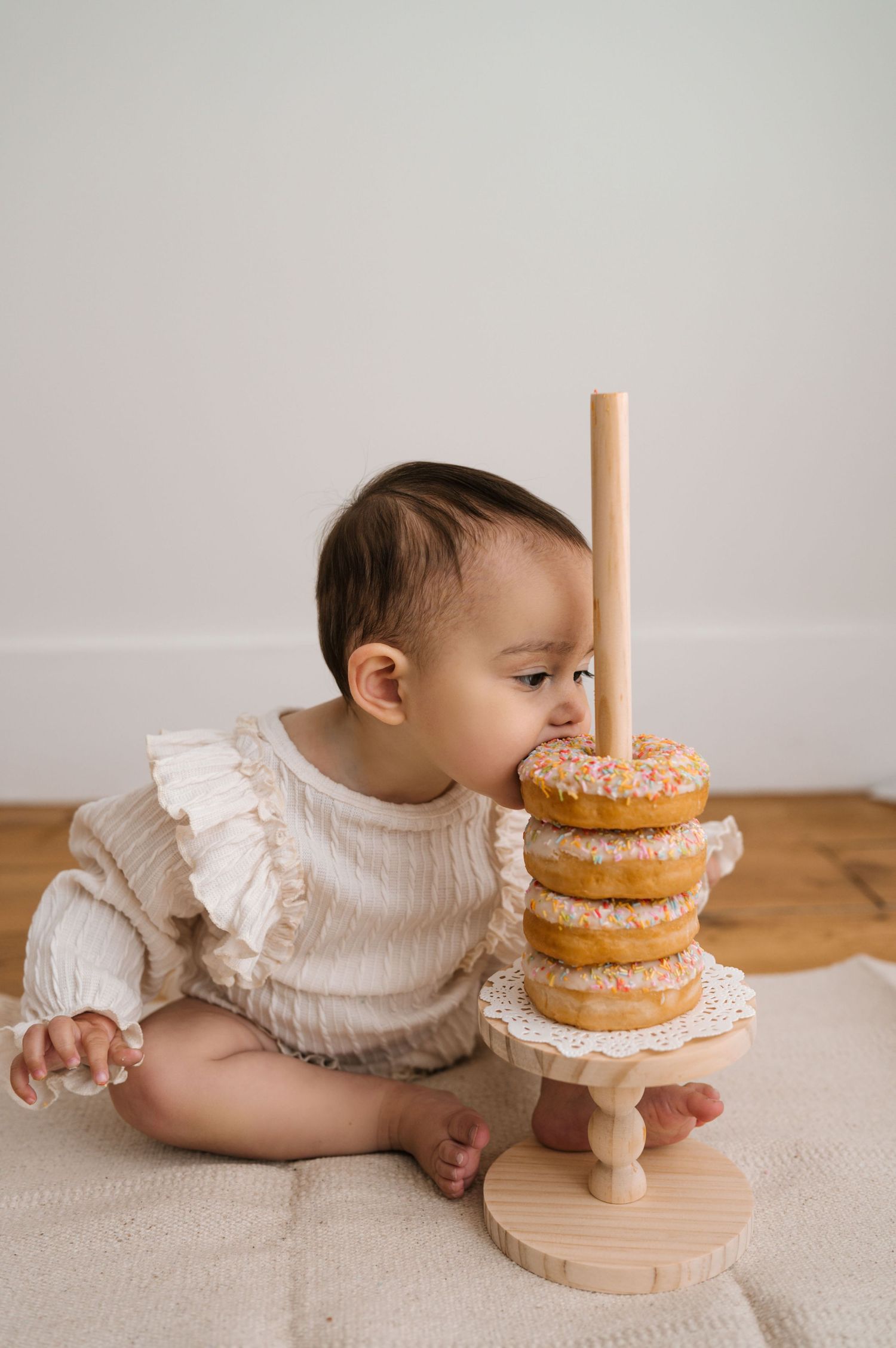 A baby in a ruffled onesie sits on a blanket, leaning in to taste a stack of sprinkled donuts on a wooden stand.