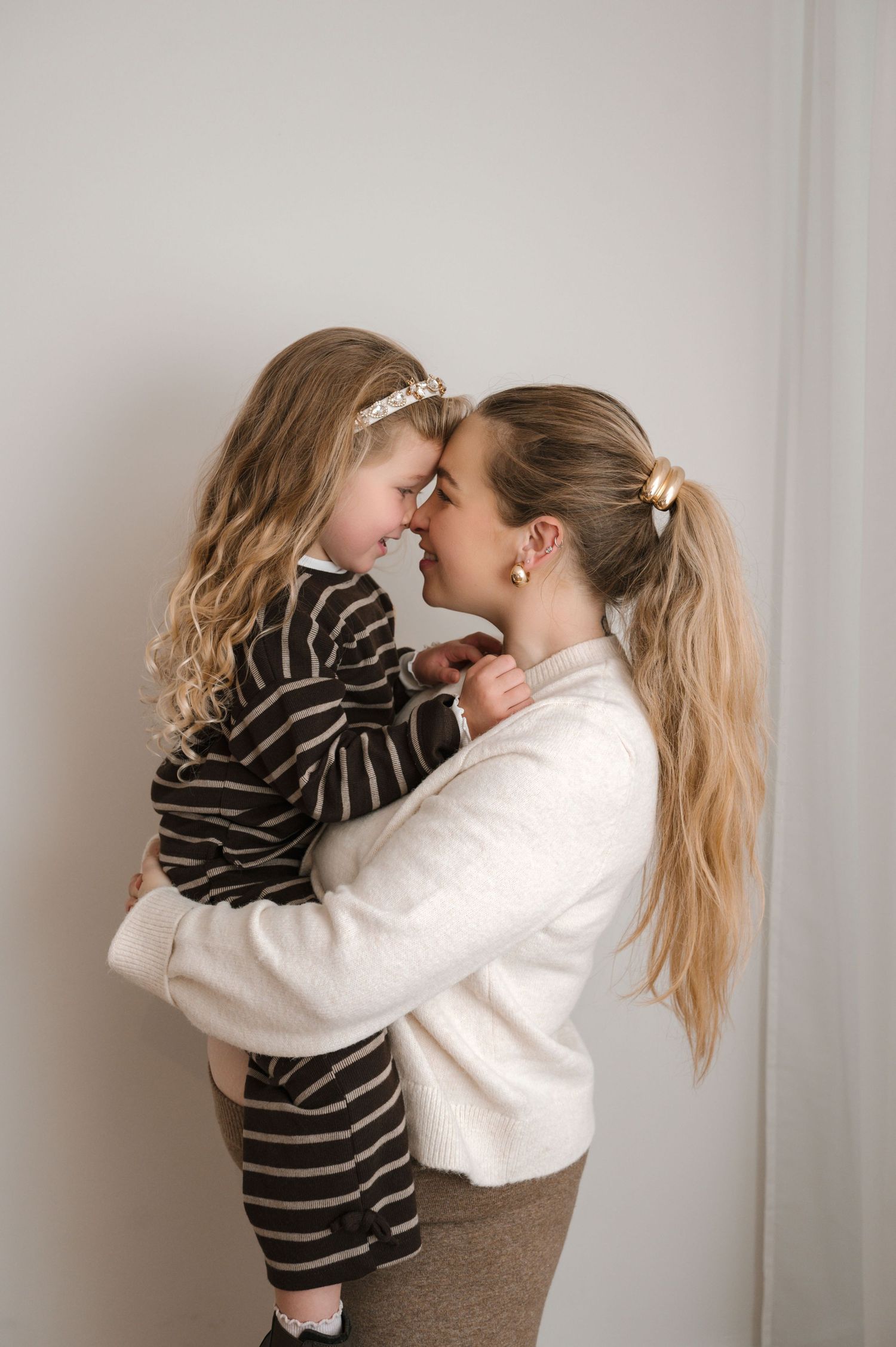 A mother holds her young blonde daughter close, touching foreheads in a tender embrace against a soft white background.