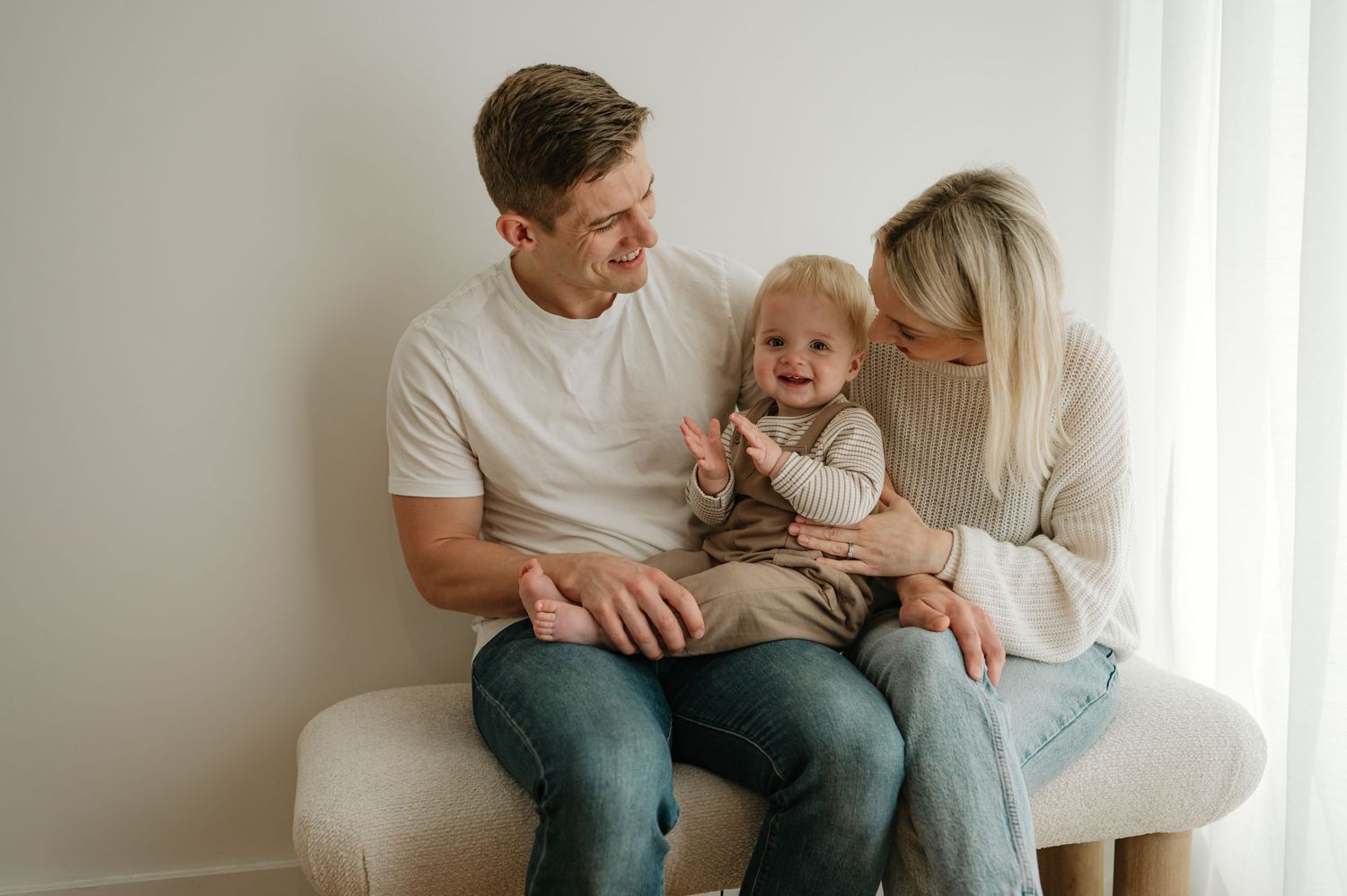 Happy young family with smiling baby sitting together on white bench in bright minimalist studio setting.