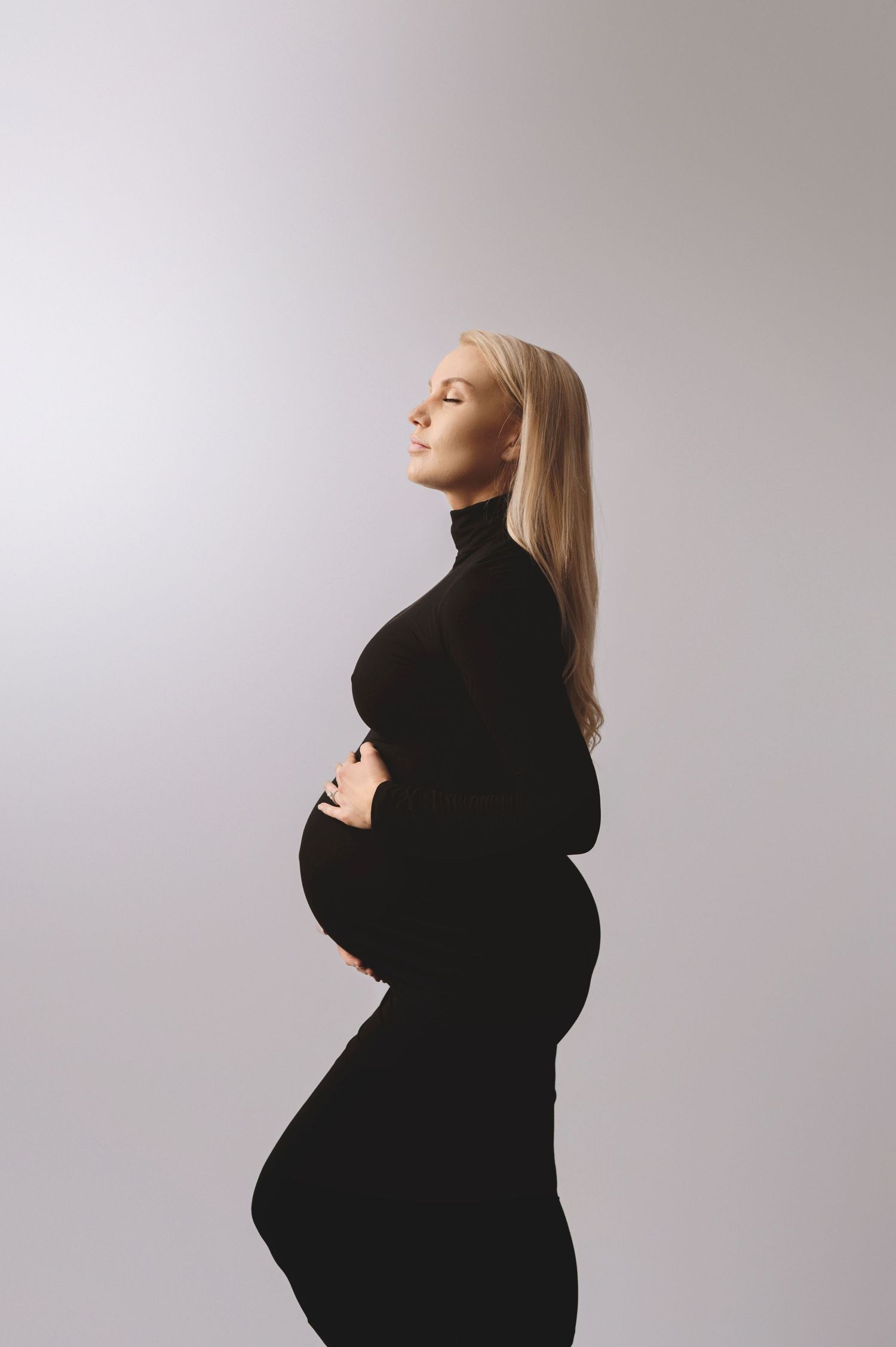Silhouetted pregnant mum in black turtleneck dress poses elegantly against light grey background in maternity photo.