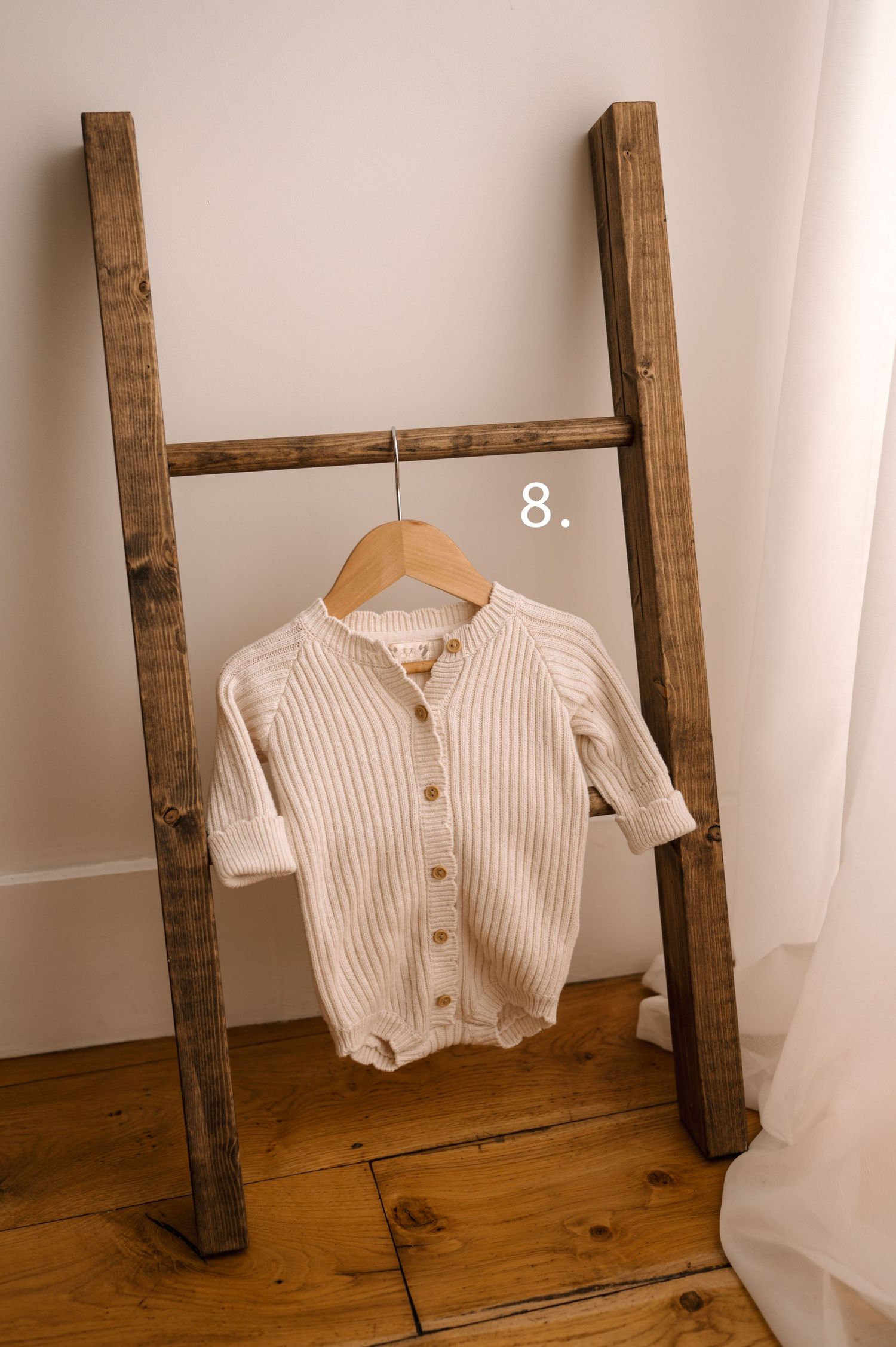 A white knitted baby cardigan sweater displayed on a rustic wooden ladder against a white wall.