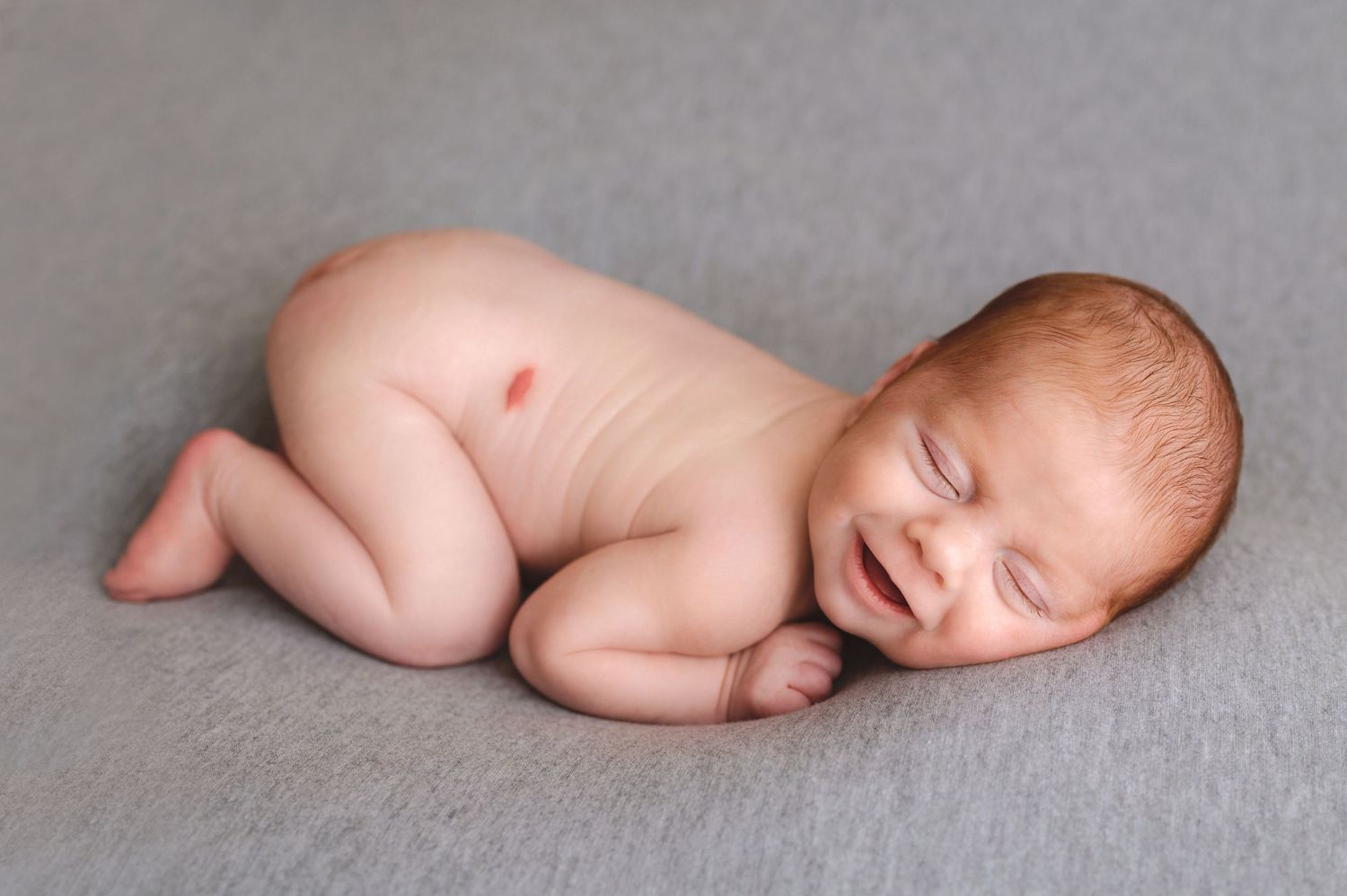 Peaceful sleeping newborn baby curled up on soft gray blanket during professional photo session.