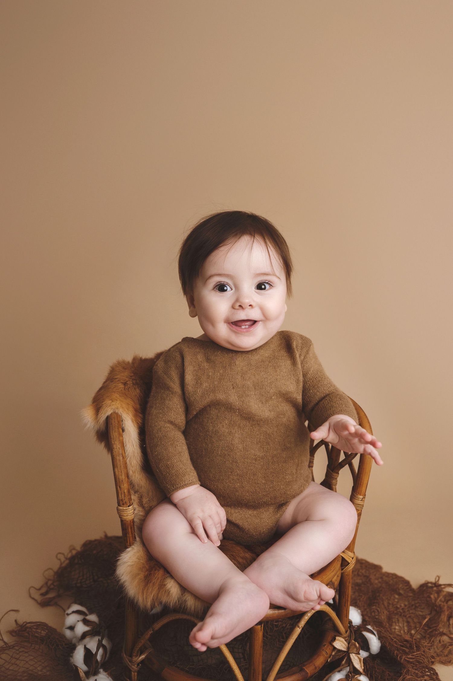 Baby in brown bodysuit smiles while sitting on wooden chair with fur lining against neutral backdrop.