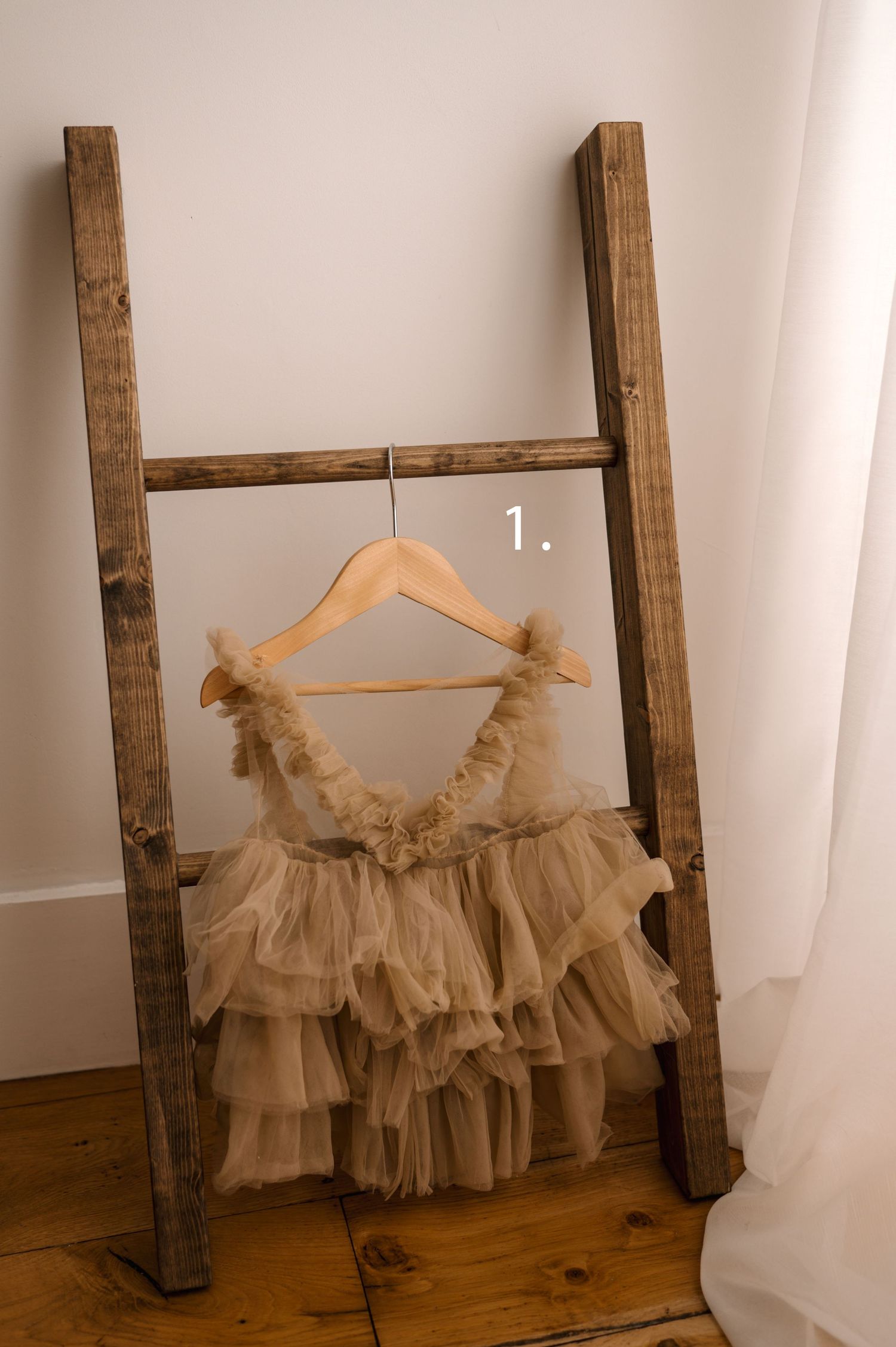 Vintage-style ruffled tulle dress displayed on a wooden hanger against a rustic ladder in soft beige tones.