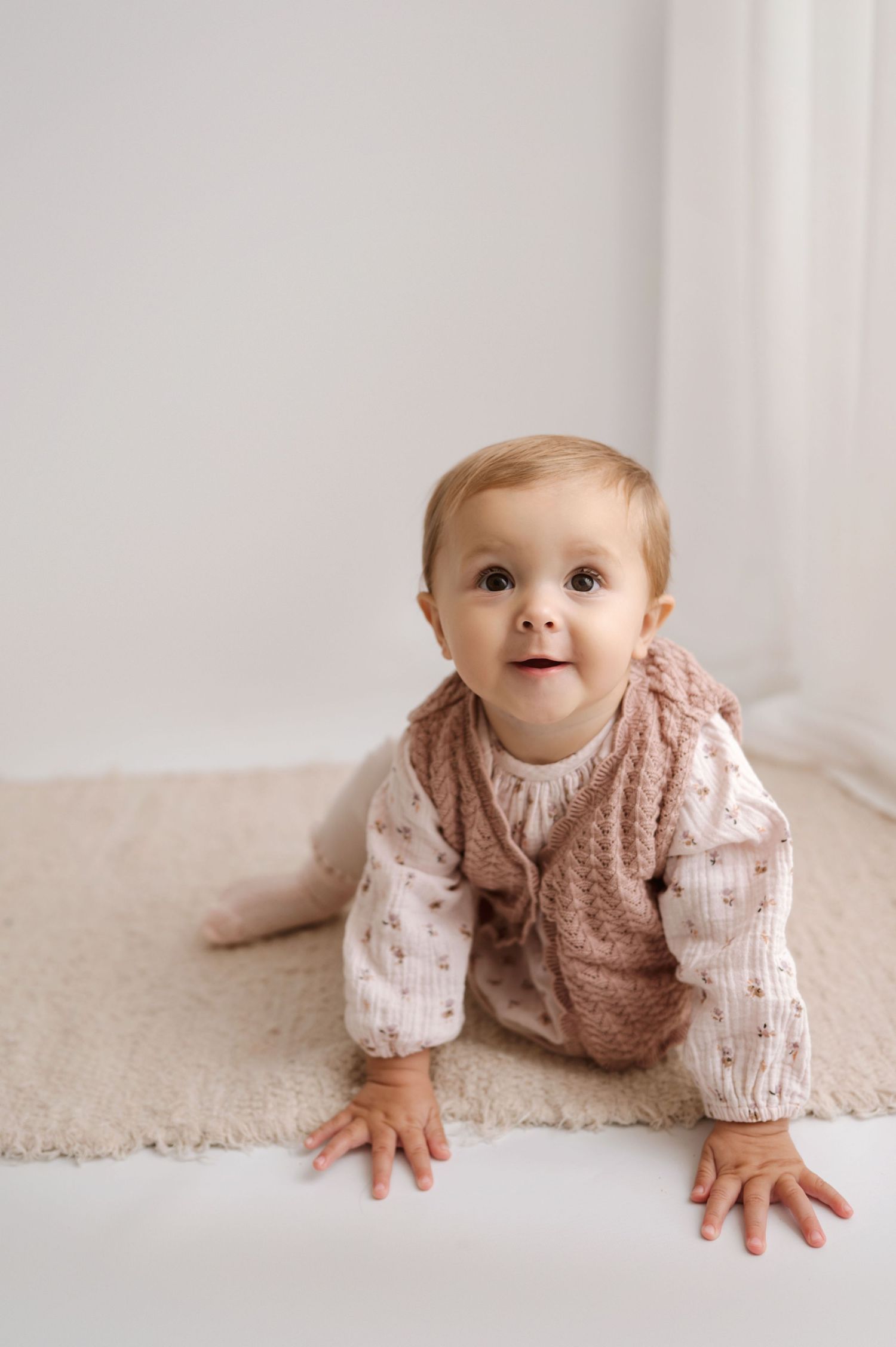 Baby wearing pink knit vest and patterned shirt crawls on beige carpet in minimalist setting.
