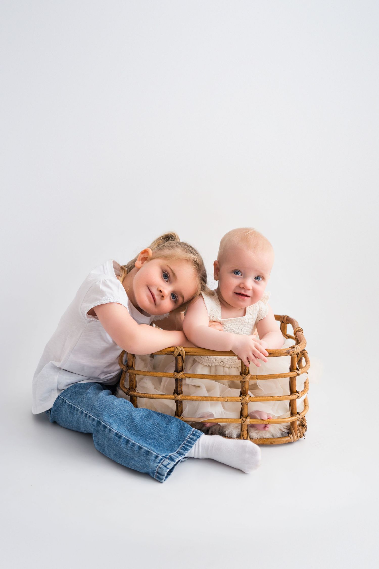 A smiling toddler girl in jeans hugs a baby sibling sitting in a wicker basket on a white background.