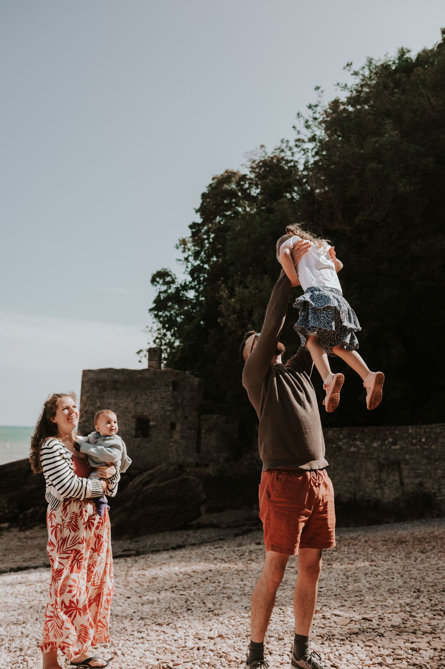 family photography enjoying a beach day as a father tosses a child in the air while mother holds a baby near coastal ruins on Elberry Cove