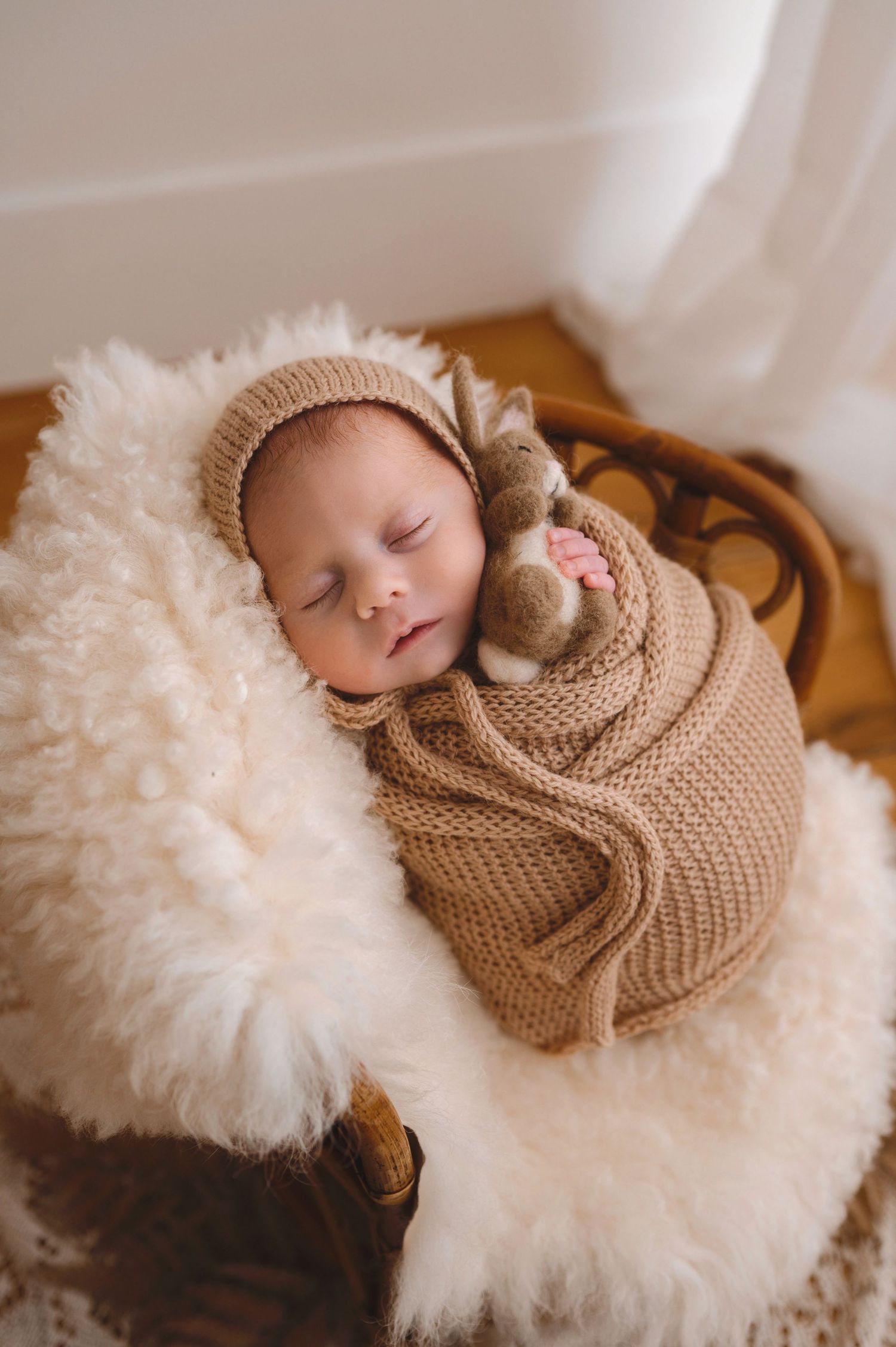 Peaceful sleeping newborn swaddled in soft beige knit wrap resting on fluffy white sheepskin blanket.