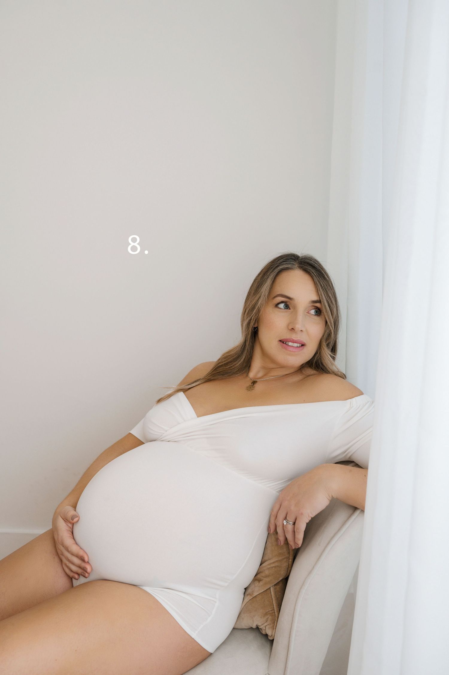 Person in white bodysuit reclining on beige couch in bright minimalist setting.