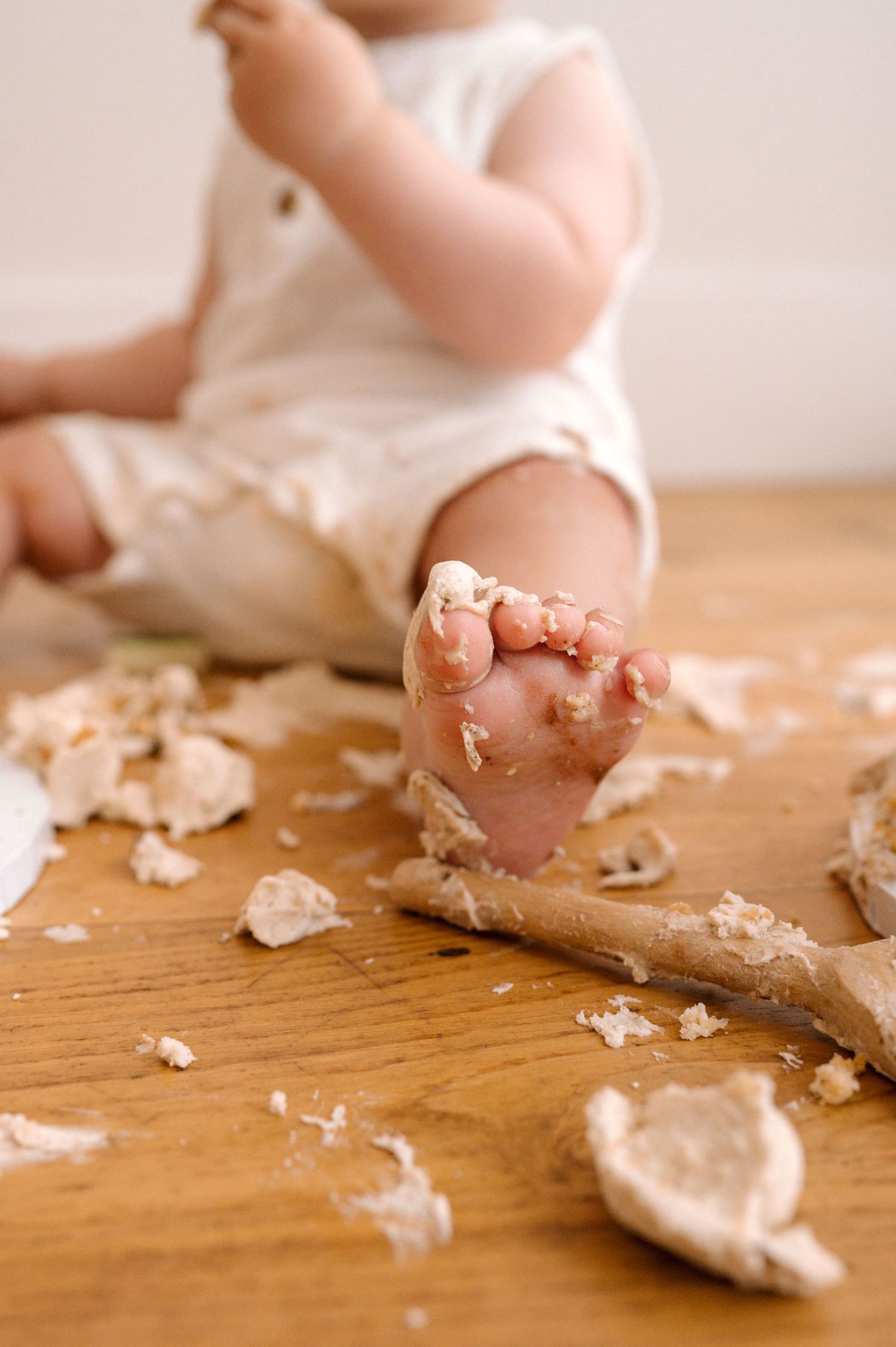 Small dusty baby feet covered in cookie crumbs on wooden floor with scattered pieces of dough and flour.