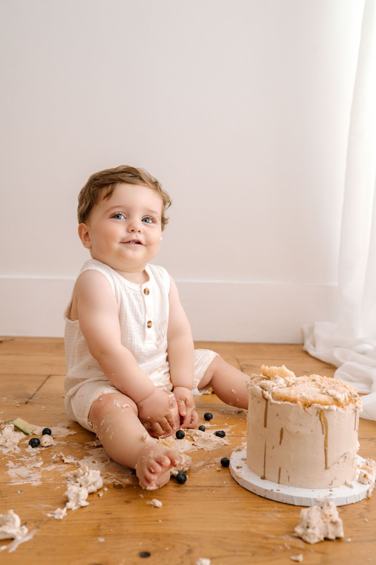 Baby in white romper sitting on wooden floor with partially destroyed birthday cake and scattered cake pieces.